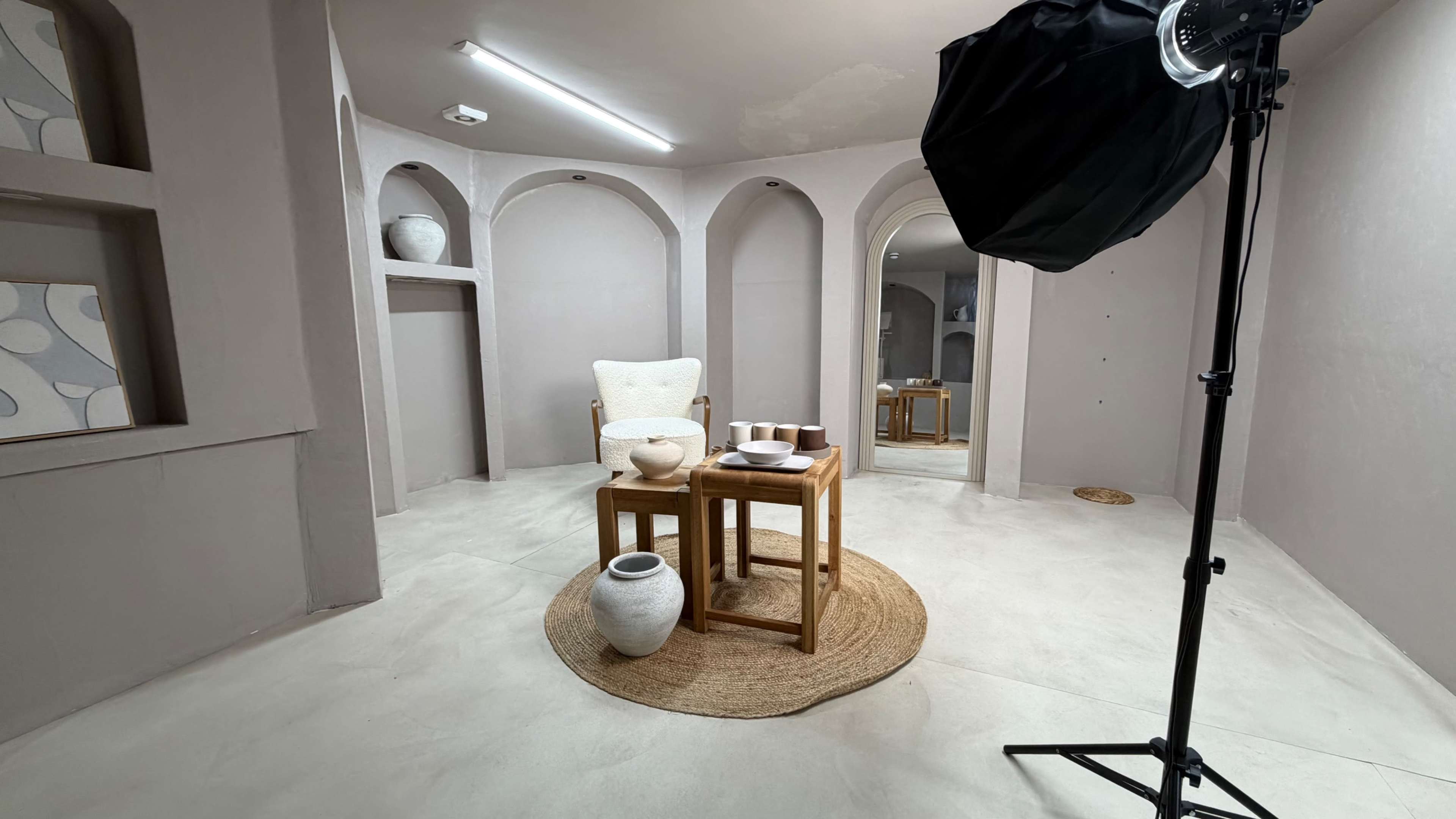 The image shows a styled interior space featuring a wooden table and chair, pottery, and a photography light setup against a minimalist backdrop with arches.