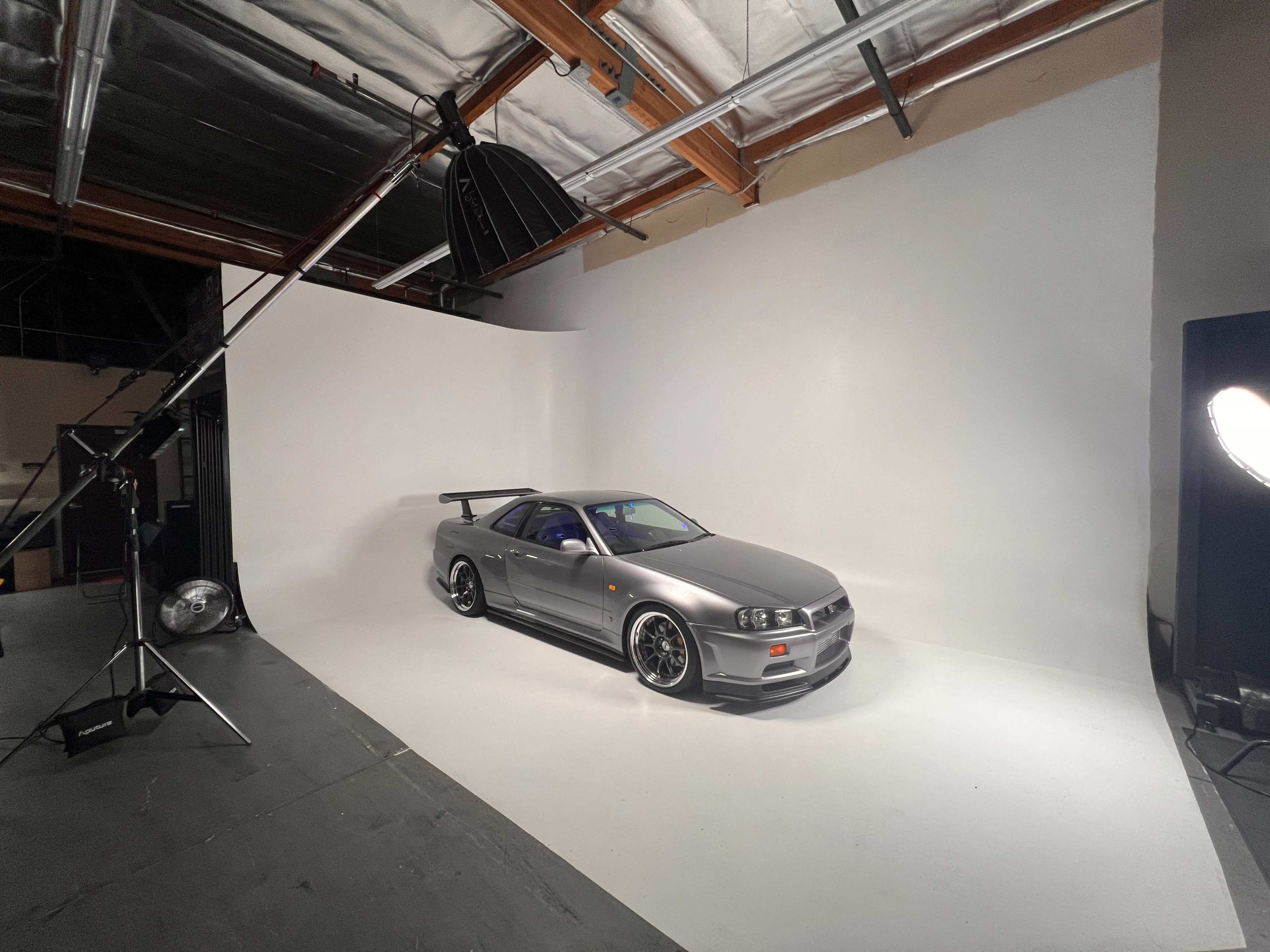 A silver sports car with a large rear wing is positioned on a seamless white backdrop in a photography studio.