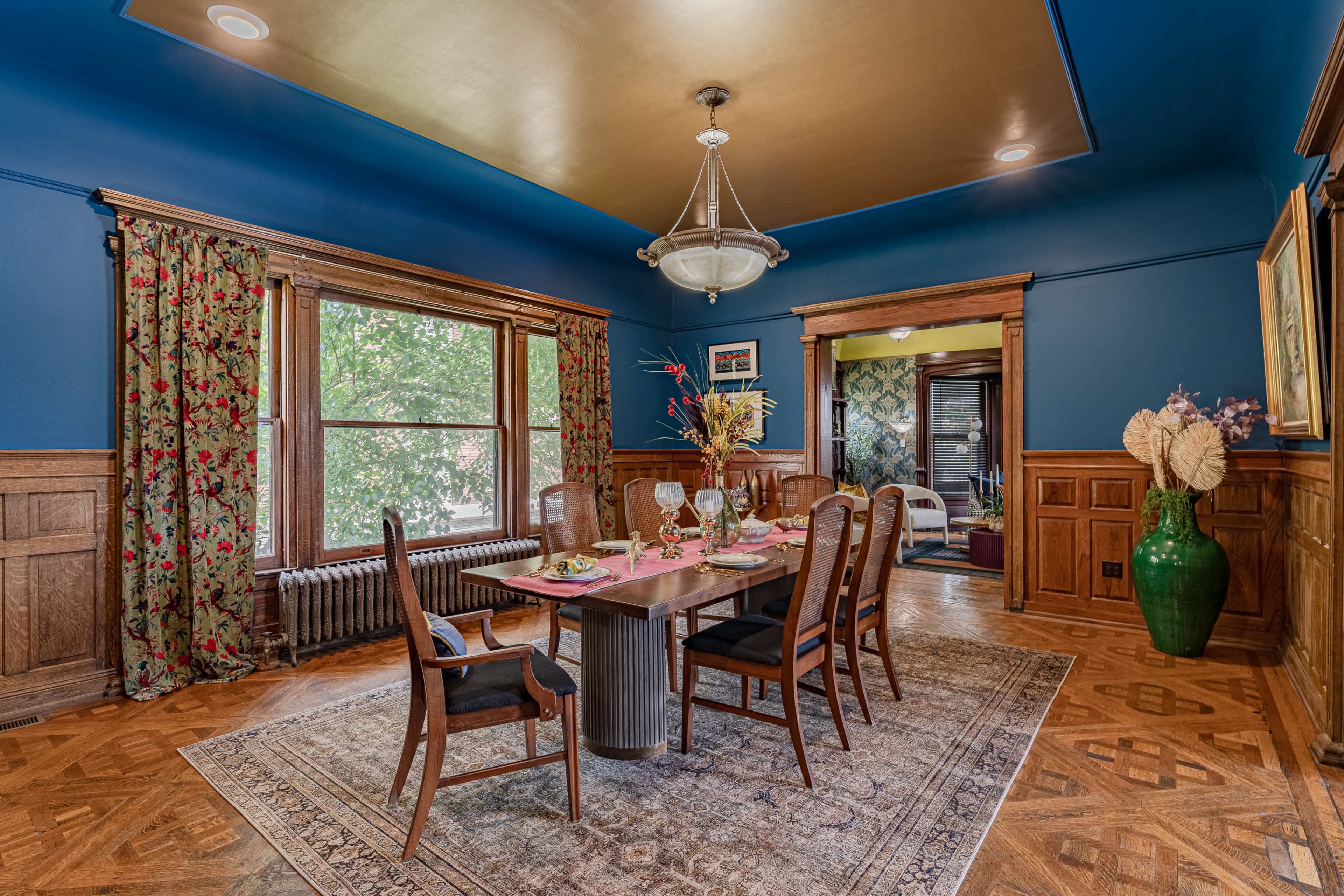 The dining room features a wooden table set with glassware, surrounded by chairs, beneath a light fixture and blue-painted walls with floral curtains.