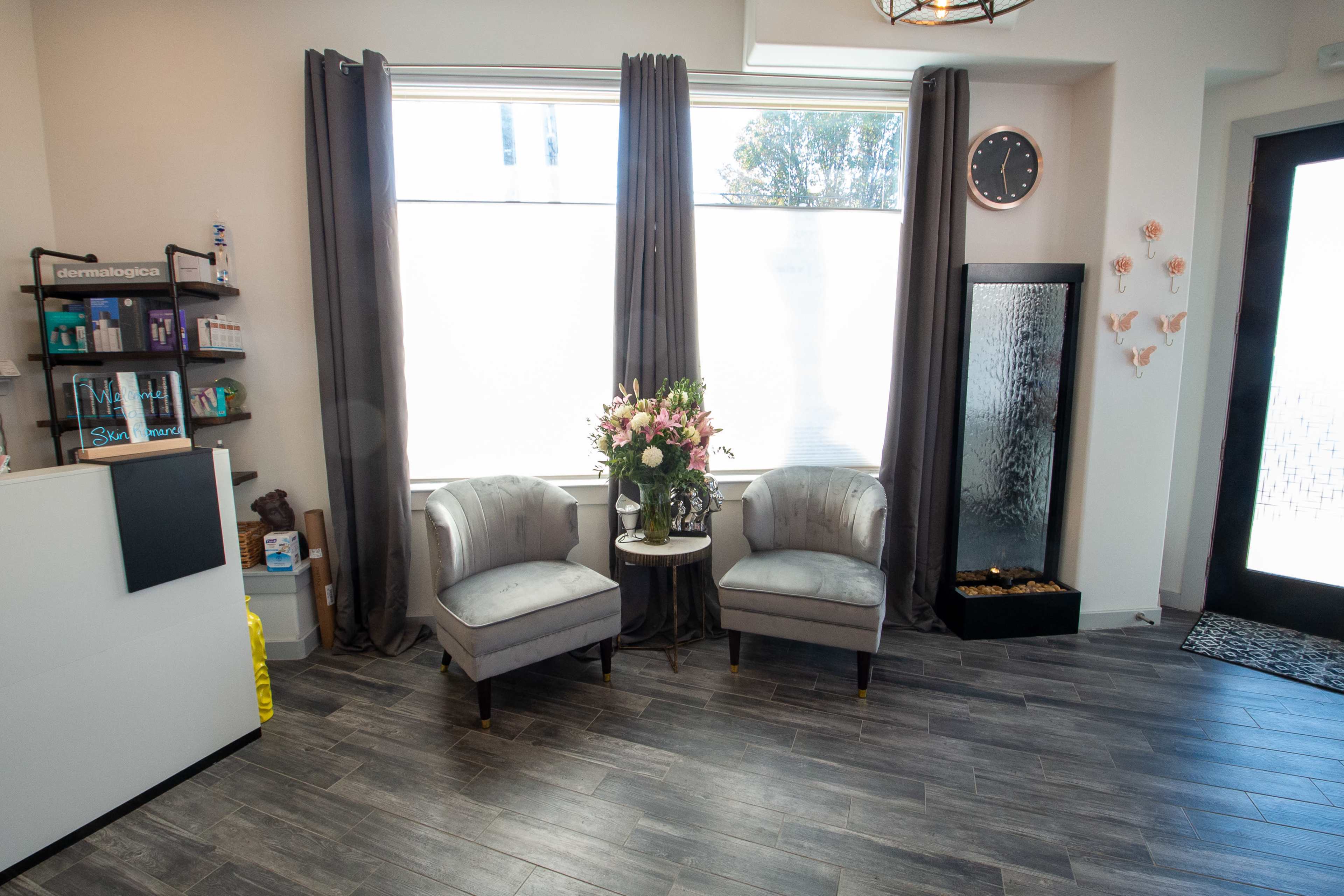 A skincare clinic reception area with two gray chairs and a small table decorated with a flower arrangement, alongside a bookshelf and a wall-mounted water feature.