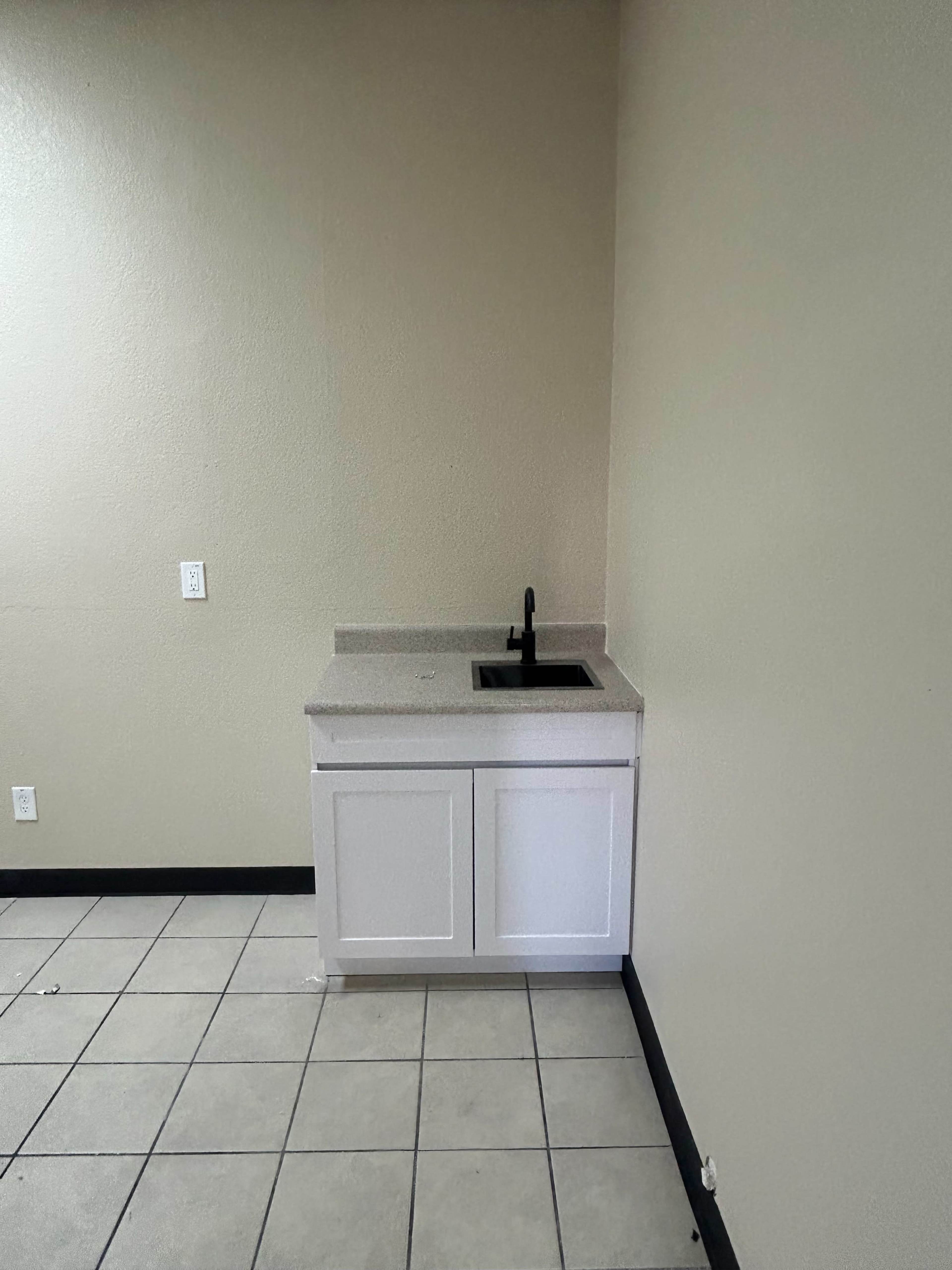 The image shows a small kitchen area featuring a countertop with a black sink and white cabinet underneath, set against a plain wall.