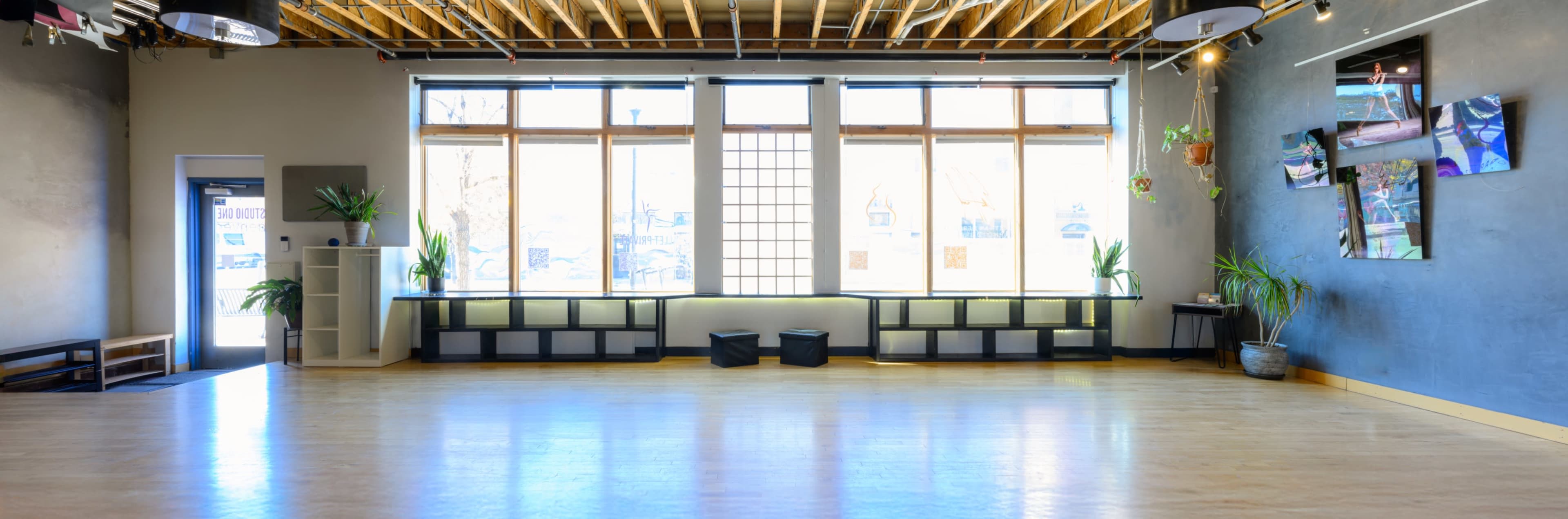 The image shows a spacious, well-lit interior room with large windows, wooden flooring, and minimalistic decor featuring plants and wall-mounted photographs.