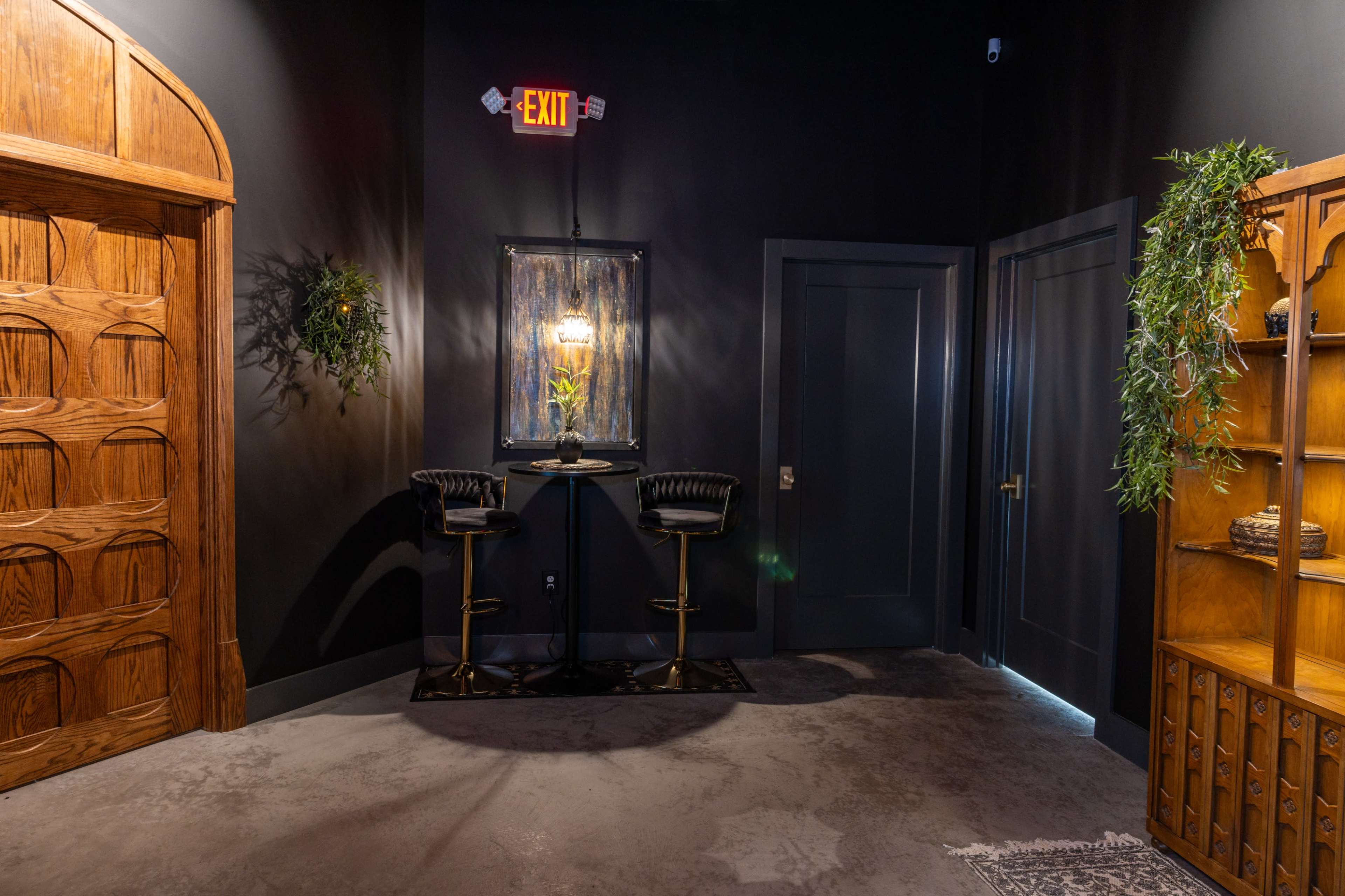 A darkly painted room with a wooden door, a small round bar table with two black stools, and a decorative mirror on the wall.