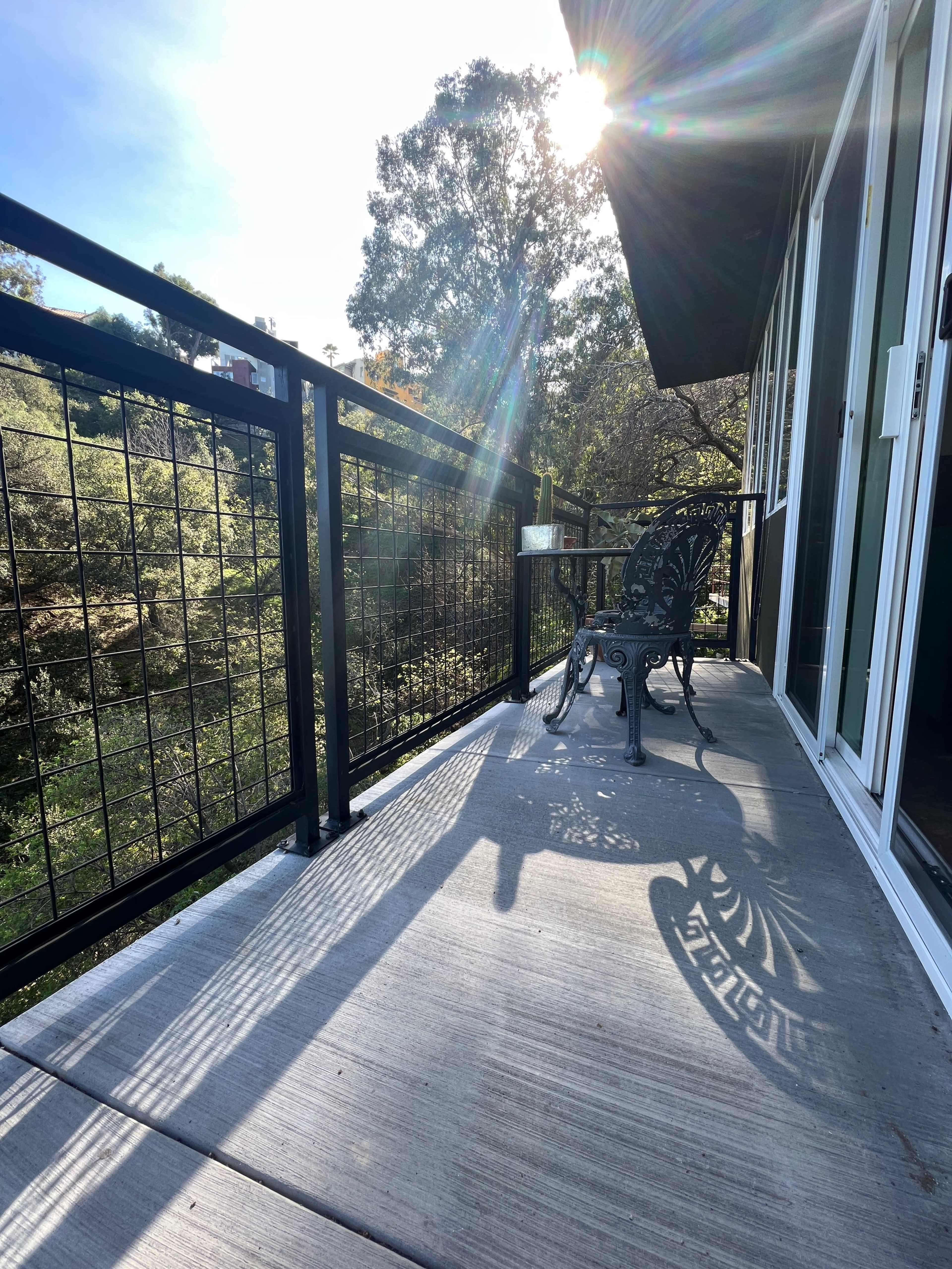 A sunlit balcony features a wrought-iron chair casting a shadow on a concrete surface, surrounded by greenery.