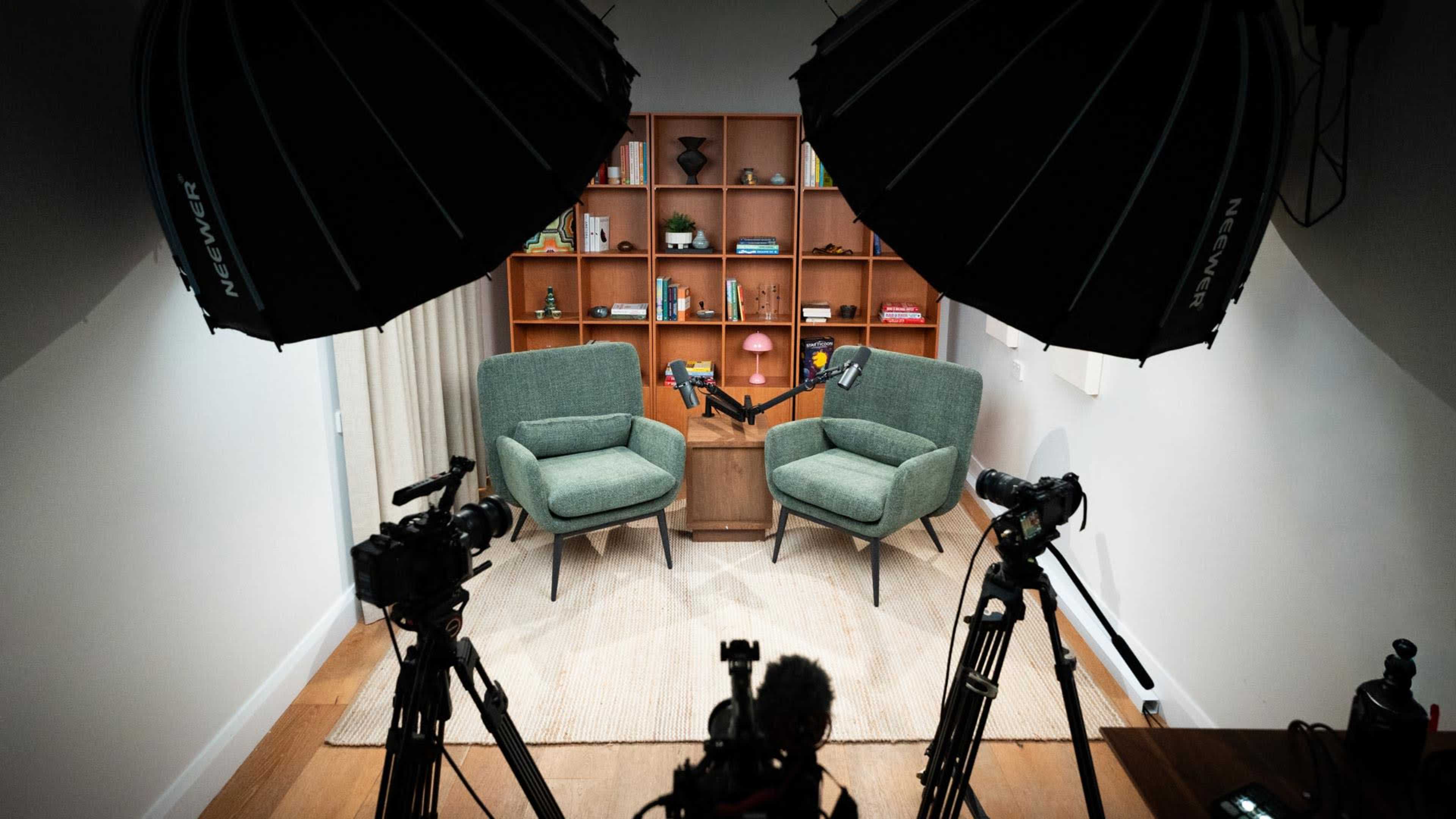The image shows two green chairs positioned in front of a wooden shelf filled with various items, with two camera setups and lighting equipment facing the chairs.
