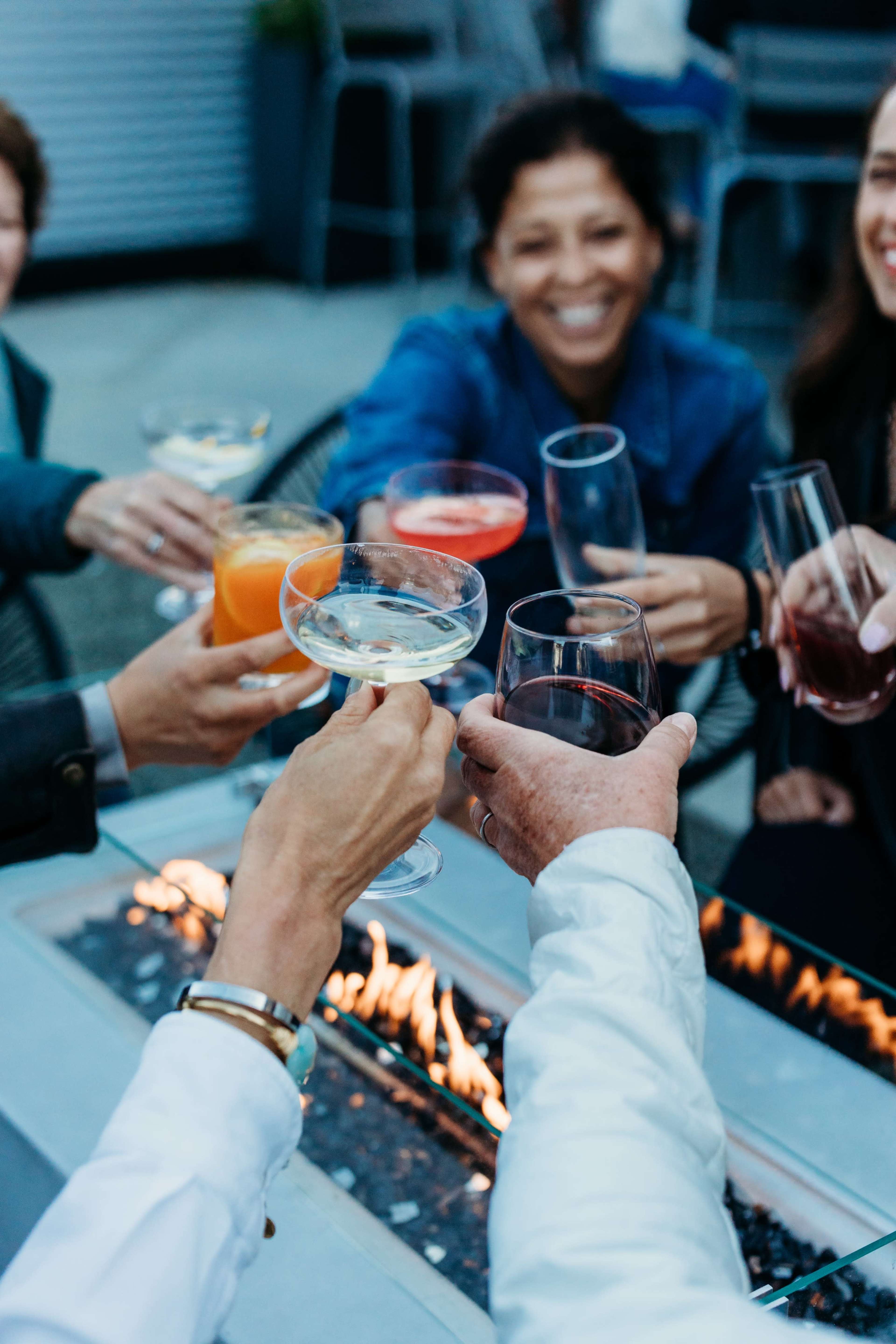 A group of people cheers with various drinks around a fire pit on an outdoor patio.