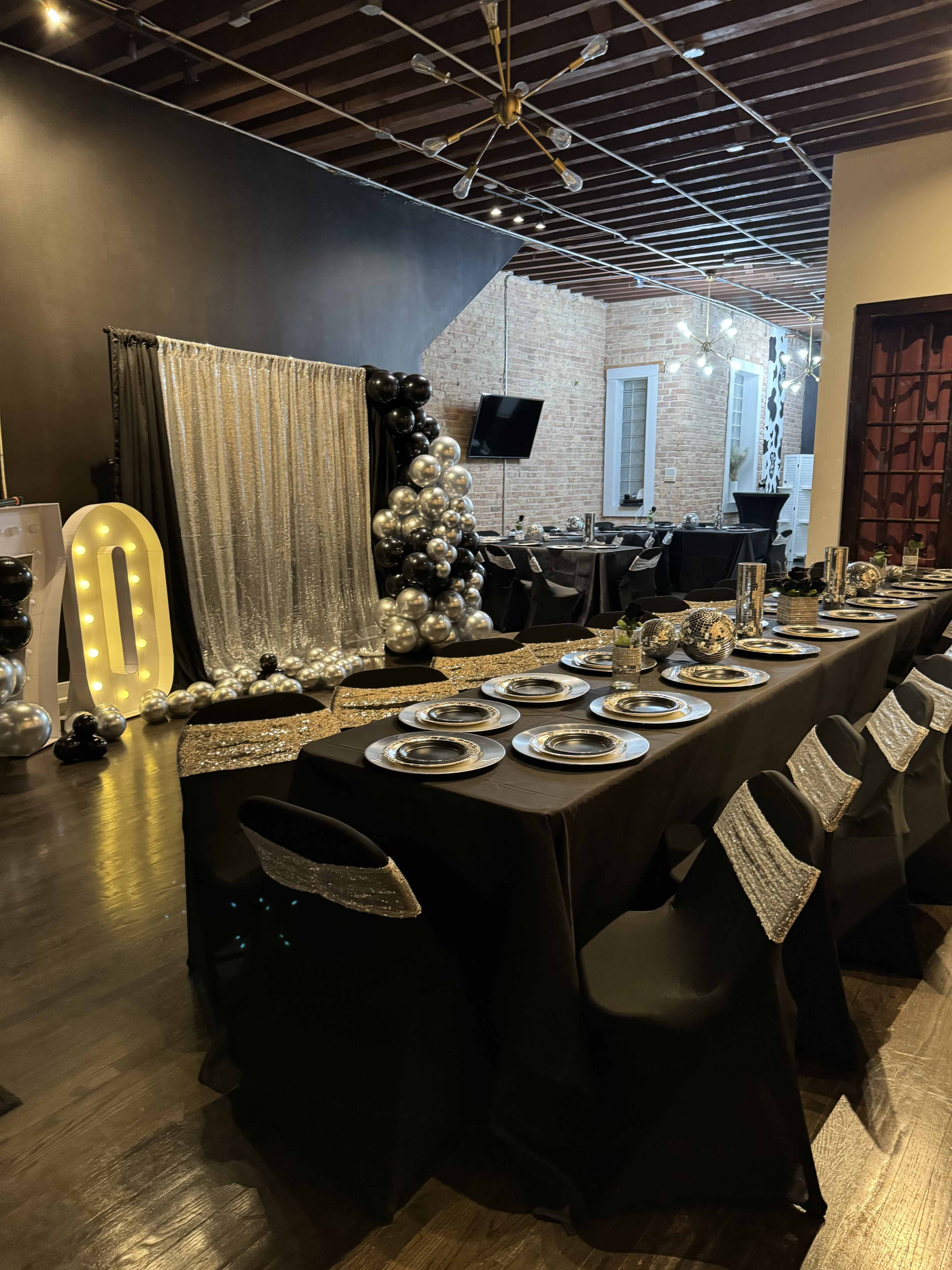 A decorated dining area with a long black table set with silver plates, surrounded by black chairs and adorned with metallic balloons.