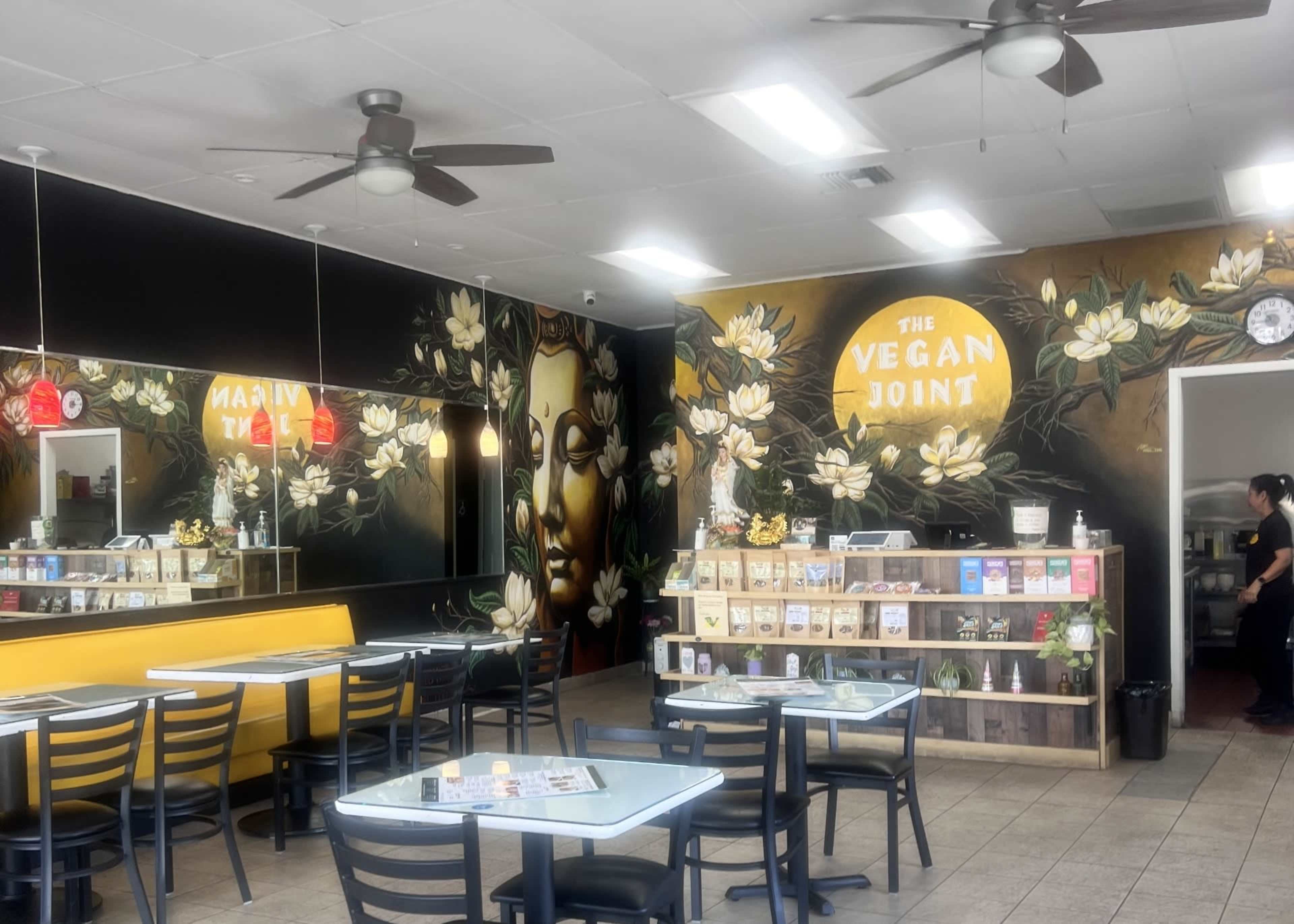 The image shows the interior of a restaurant called "The Vegan Joint" with floral murals on the walls, a service counter with menu items displayed, and tables and chairs for dining.