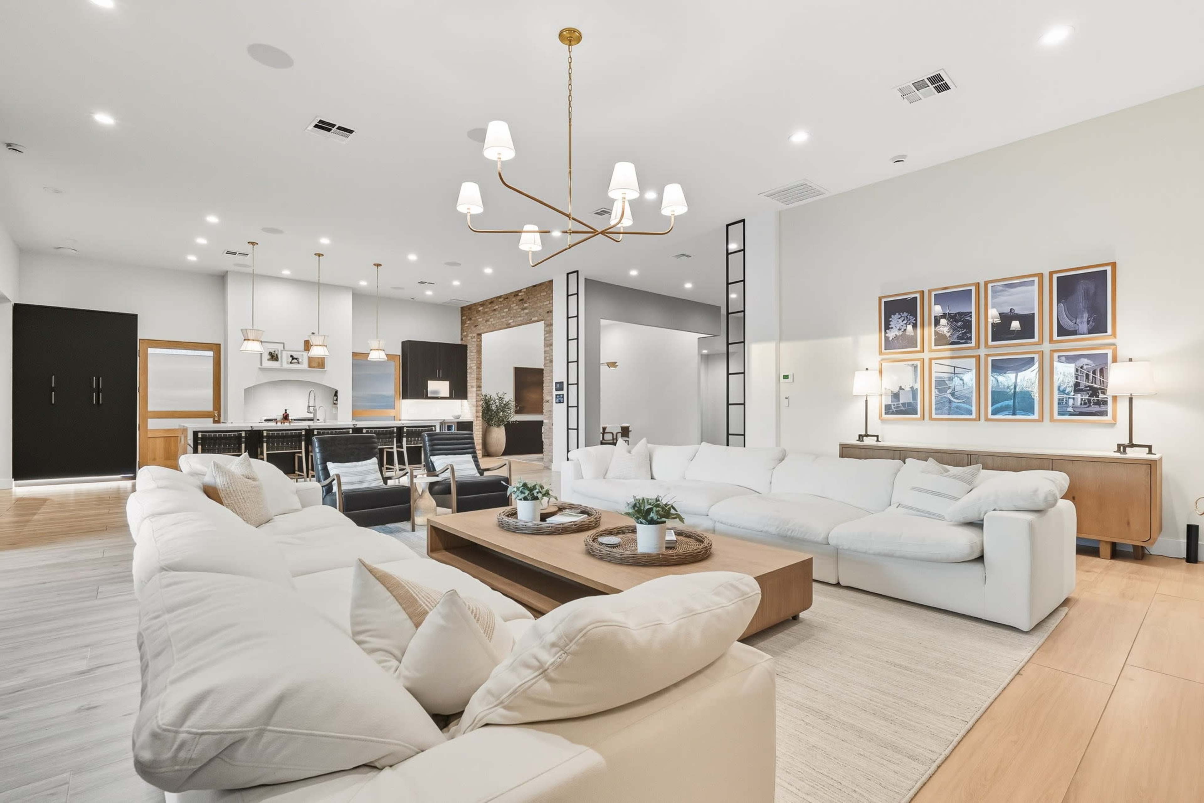 A spacious living room features a large sectional sofa, a wooden coffee table, and a modern chandelier, with an open view of a kitchen in the background.
