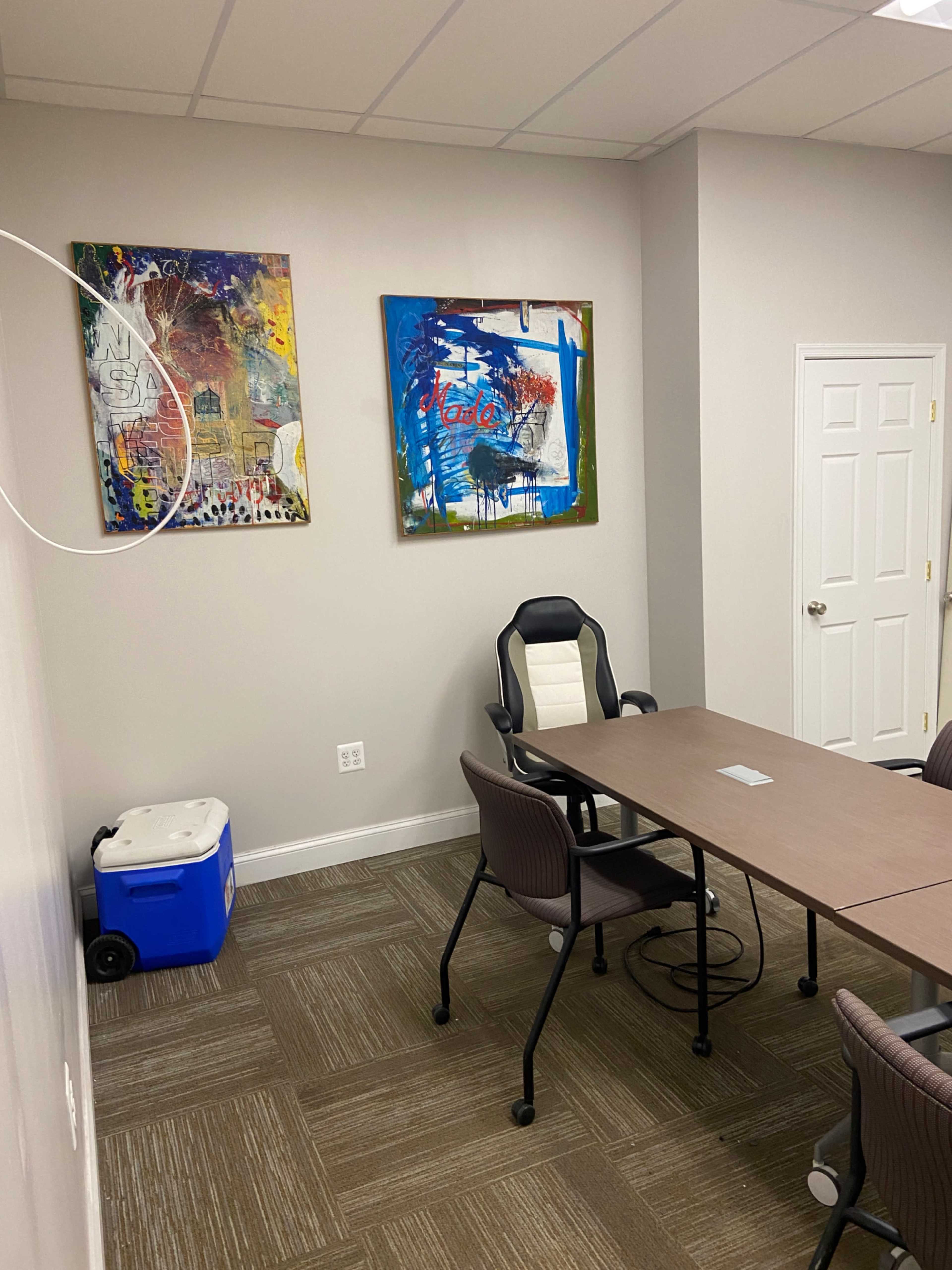 A small meeting room with a rectangular table, several chairs, two colorful paintings on the wall, and a cooler in the corner.