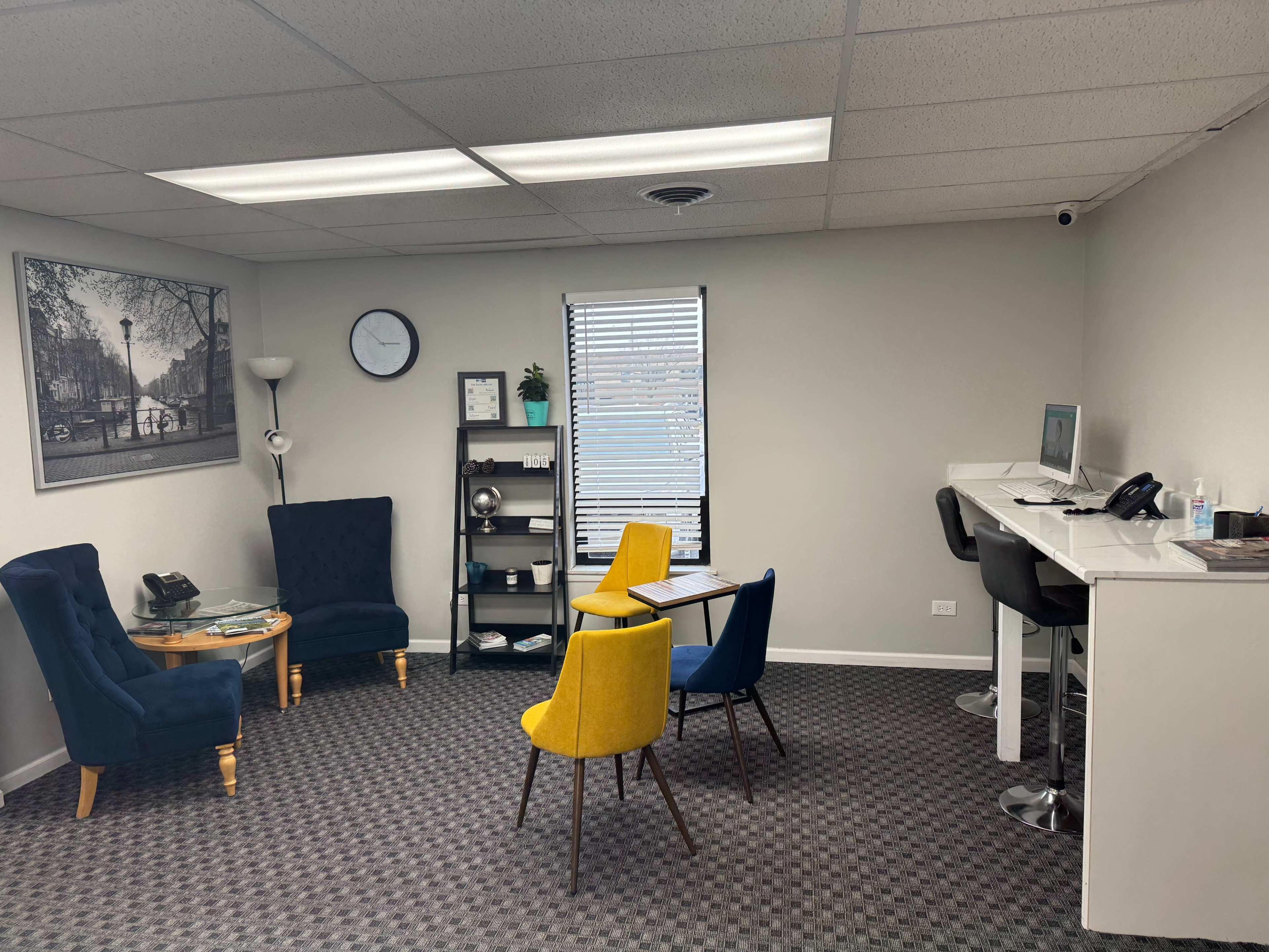 A small office space with two blue chairs, two yellow chairs, a glass table, a desk with a computer, and a wall clock.