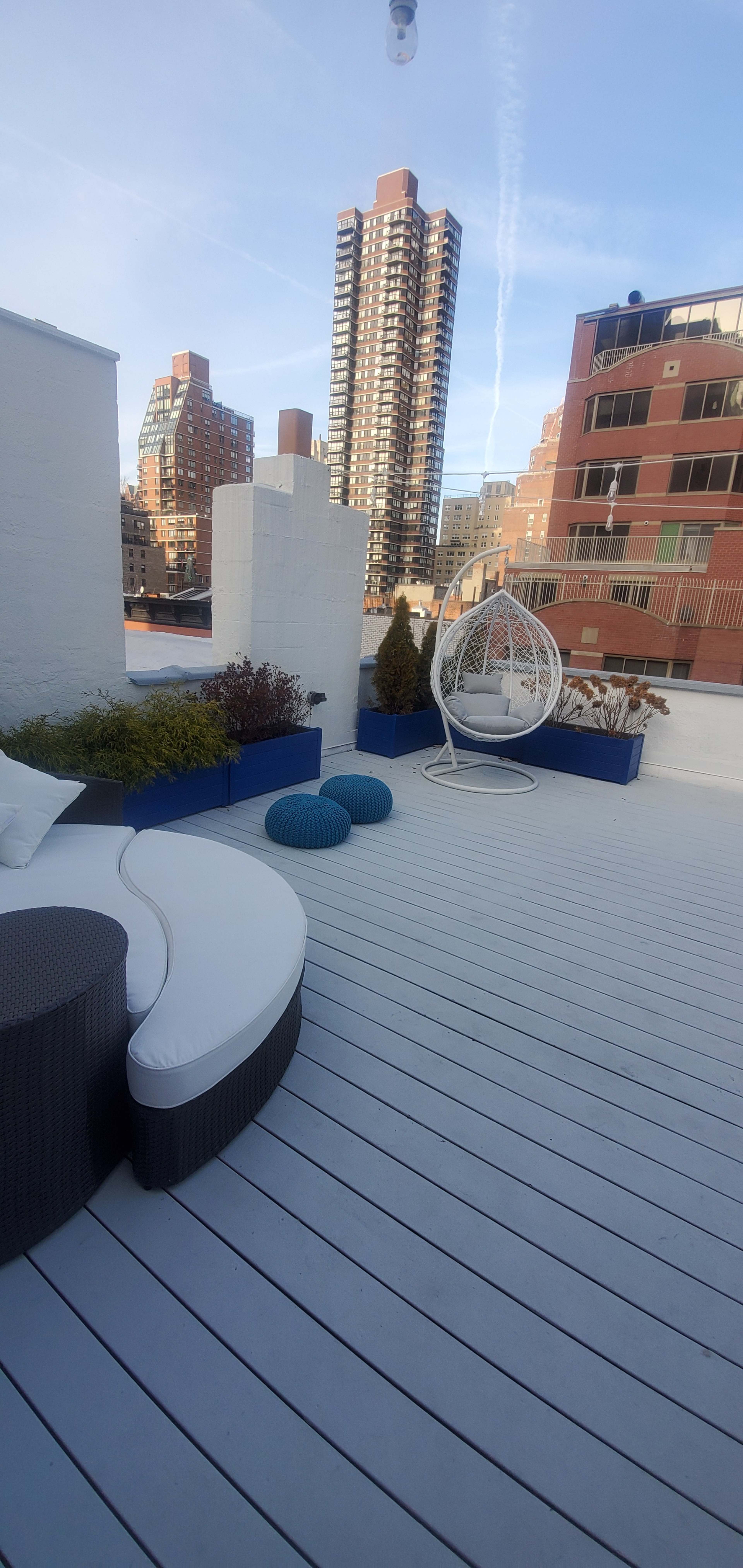 A rooftop terrace featuring a curved white couch, blue decorative poufs, and a hanging chair, with tall city buildings in the background.