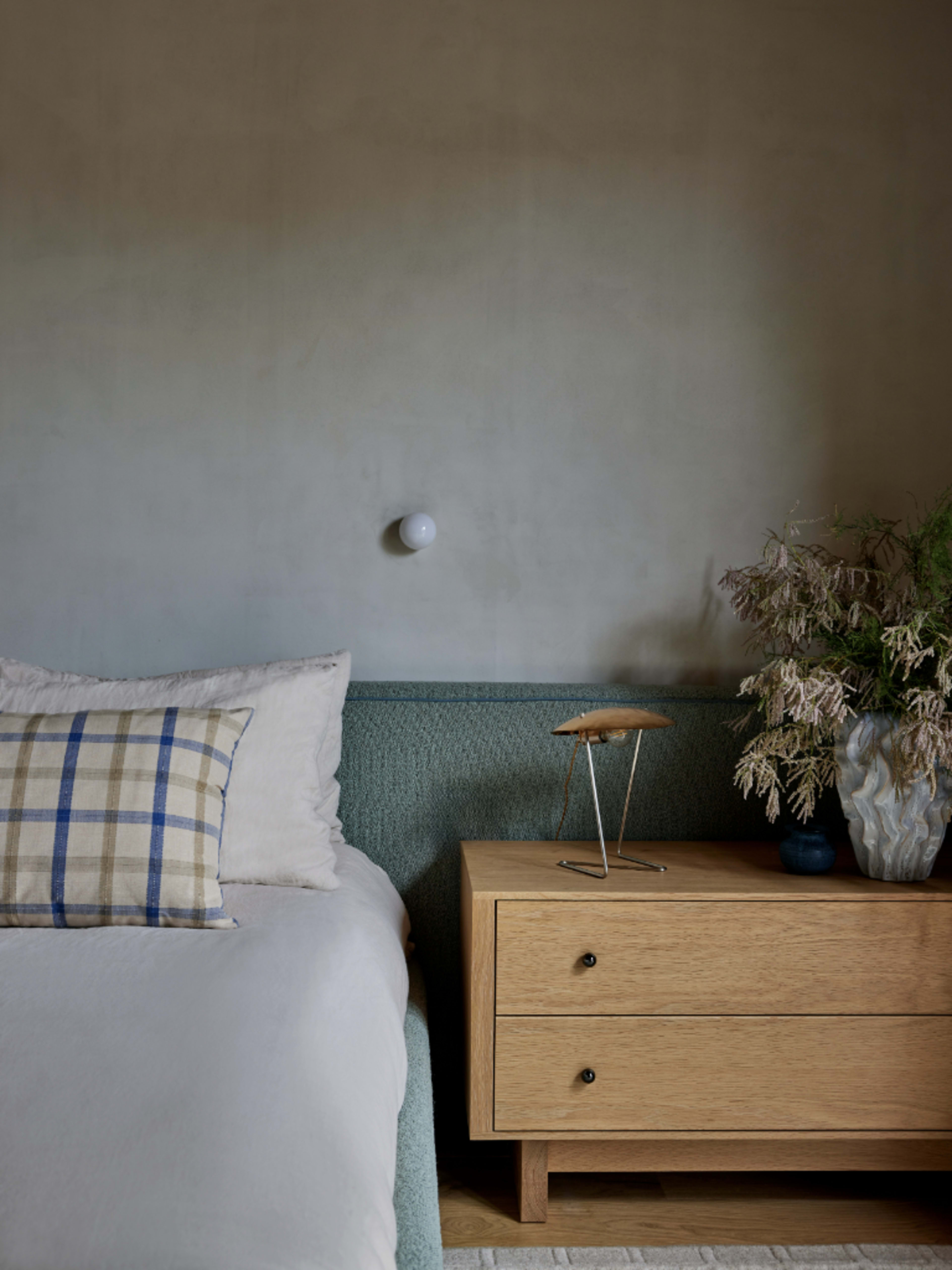 A cozy bedroom featuring a bed with a plaid pillow next to a wooden nightstand and a decorative vase with dried flowers.