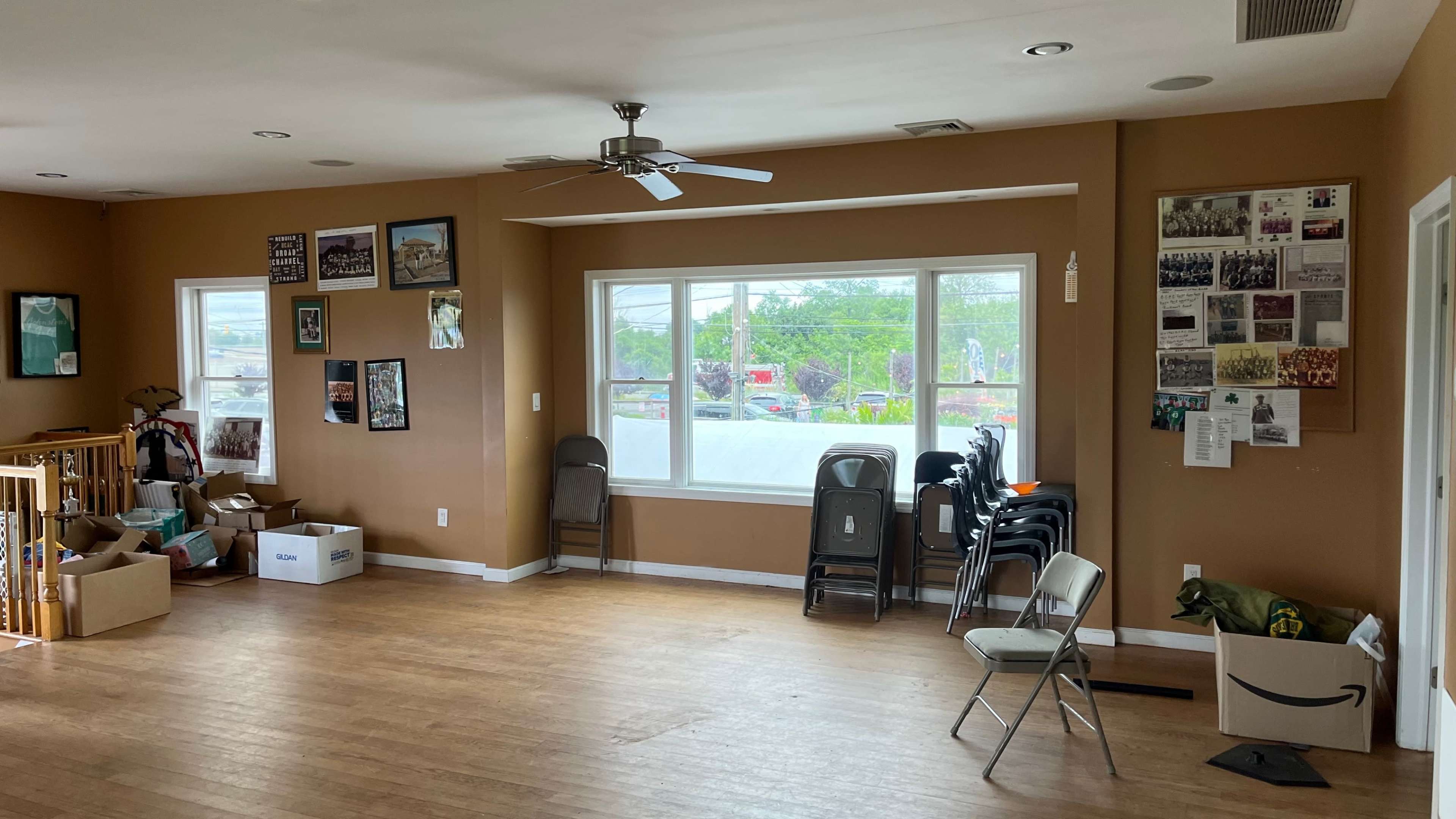 The room features wooden flooring, a ceiling fan, several chairs stacked against a wall, and various framed photos displayed on the walls.