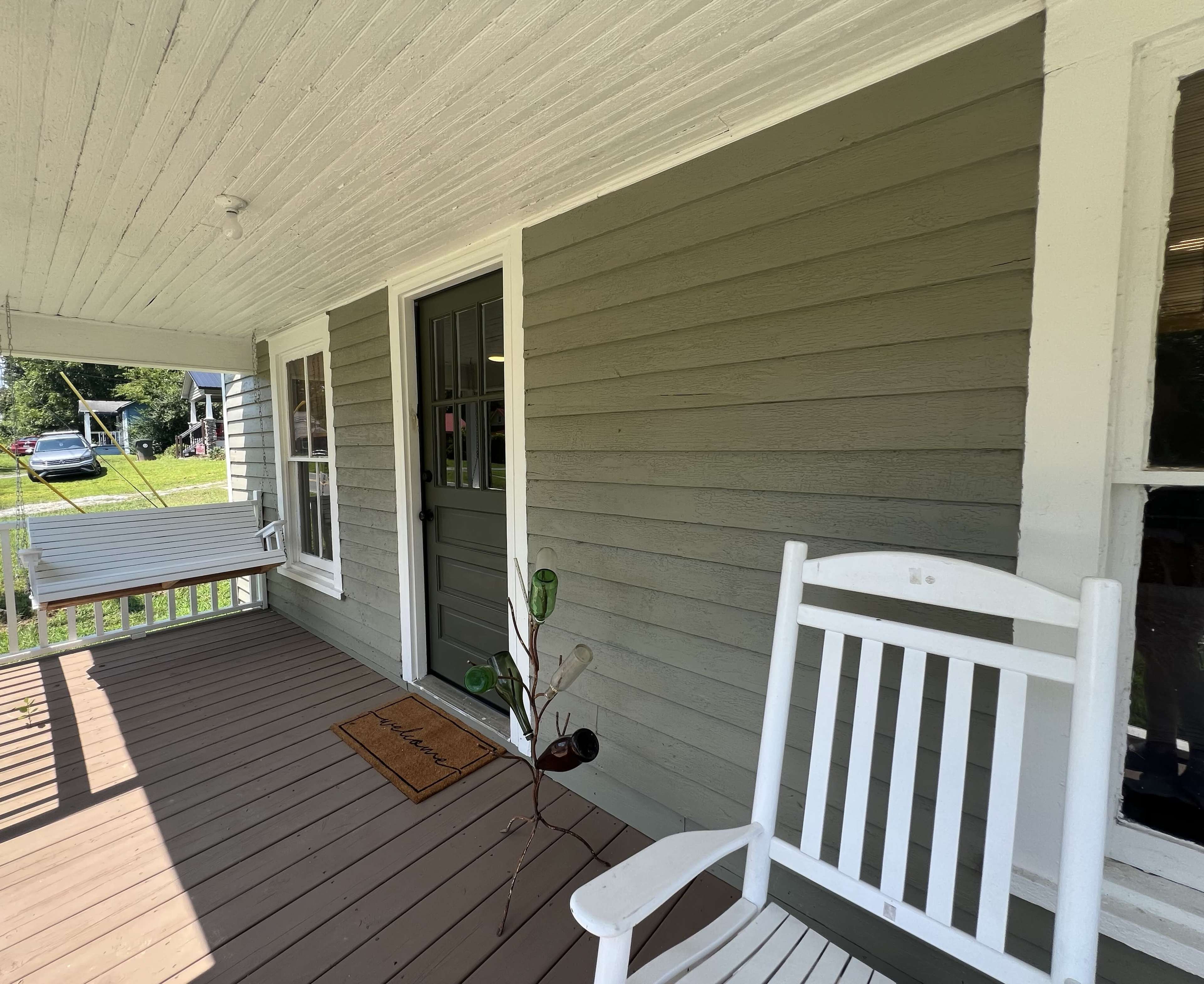 The image shows a porch area of a house featuring a white rocking chair, a welcome mat, and a green door with nearby plants.