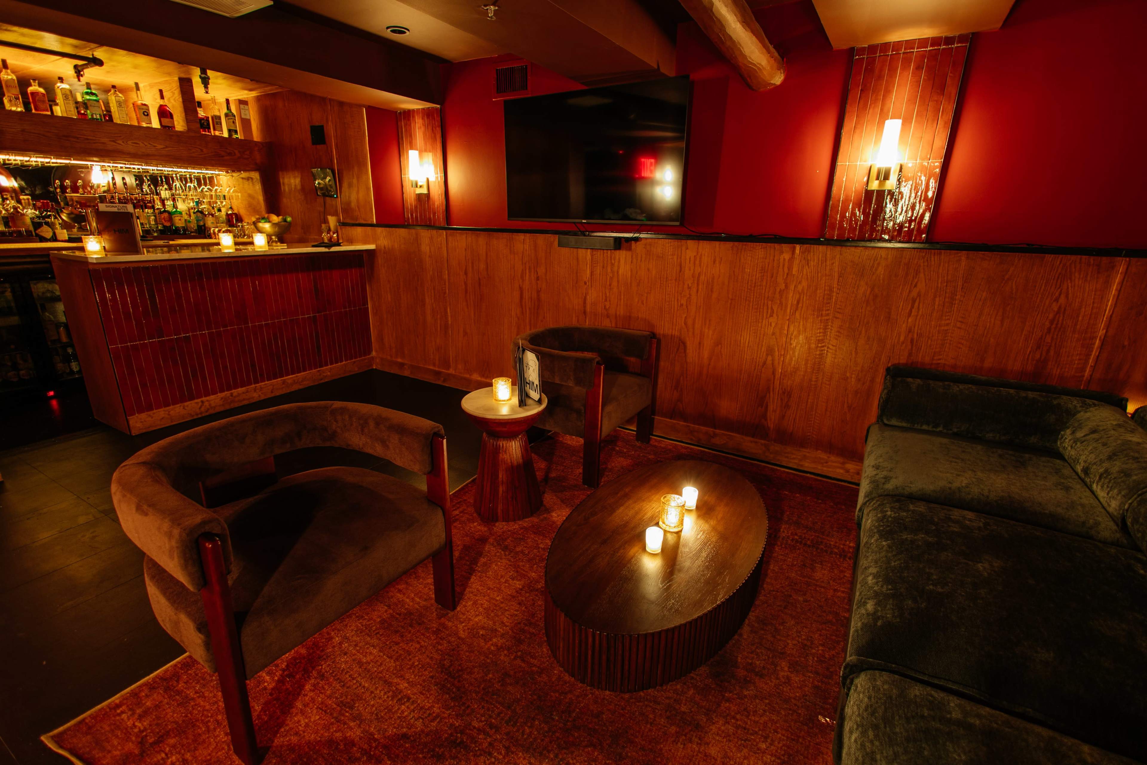 The image depicts a cozy lounge area with two chairs, a small table, and a sofa, surrounded by dim lighting and wooden accents.