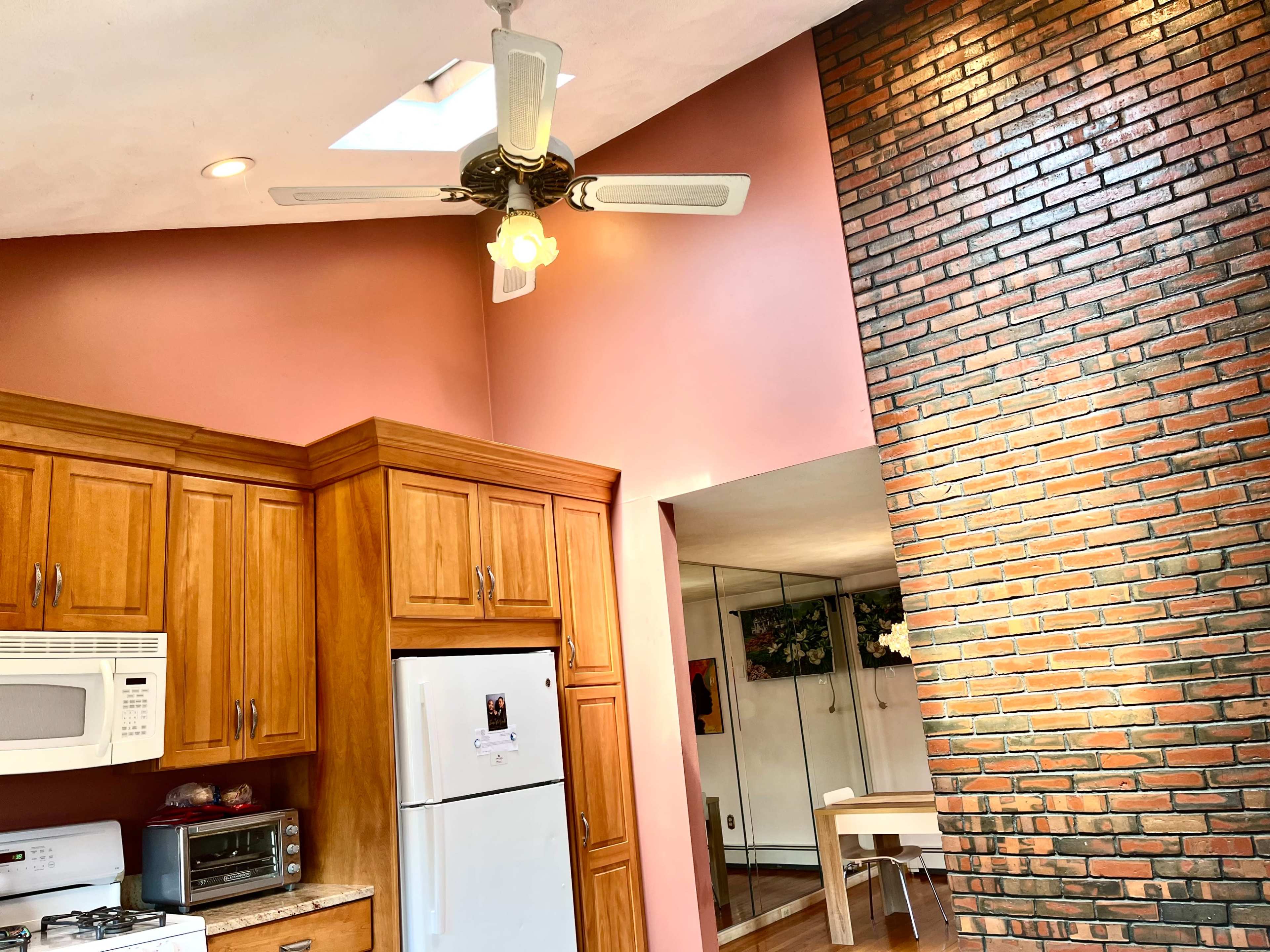 The image shows a kitchen with wooden cabinets, white appliances, a ceiling fan, and a brick wall adjacent to a dining area with a table.
