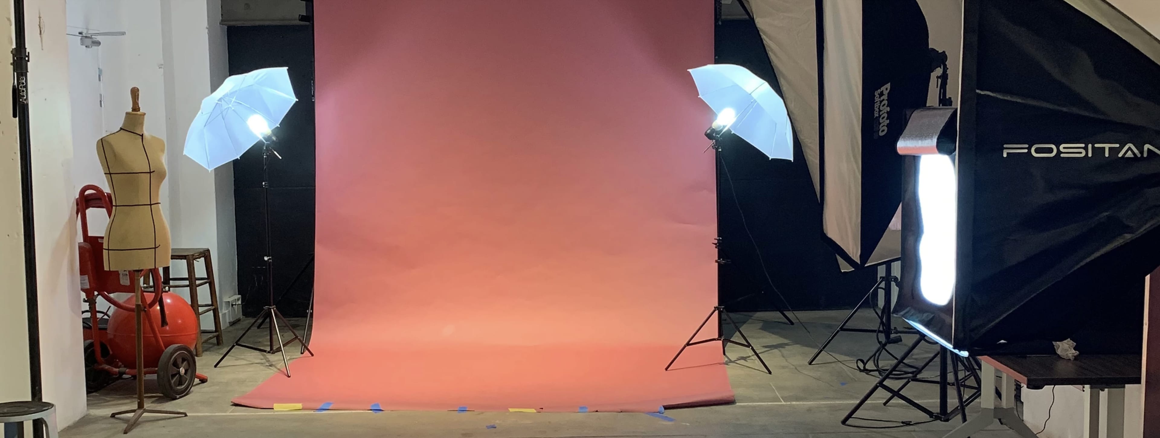 The image shows a photography studio setup with a pink backdrop, two softbox lights, and various equipment including a dress form and a chair.