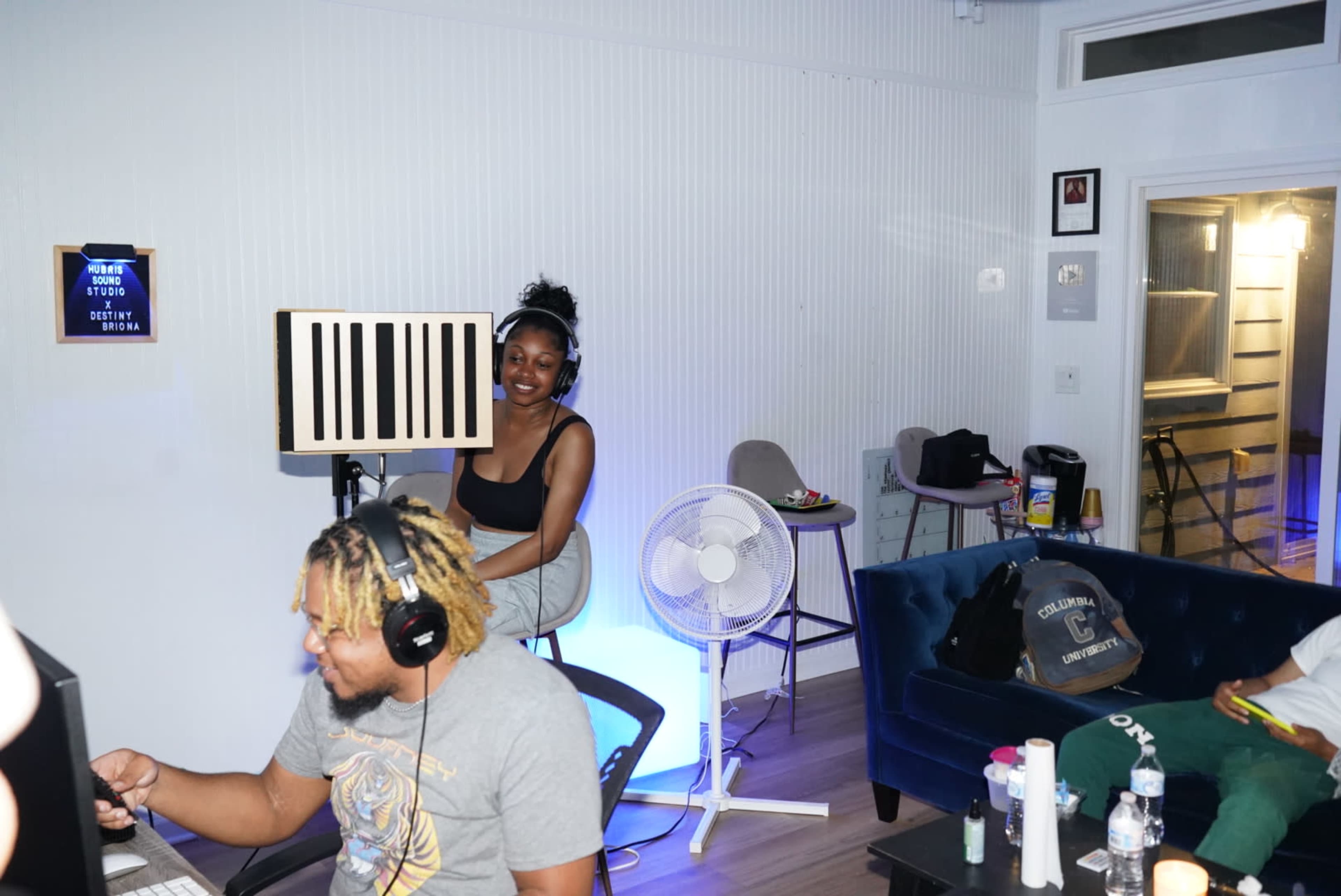 A woman stands next to a striped artwork while a group of people sits at a table with computers in a room featuring modern furniture and a fan.