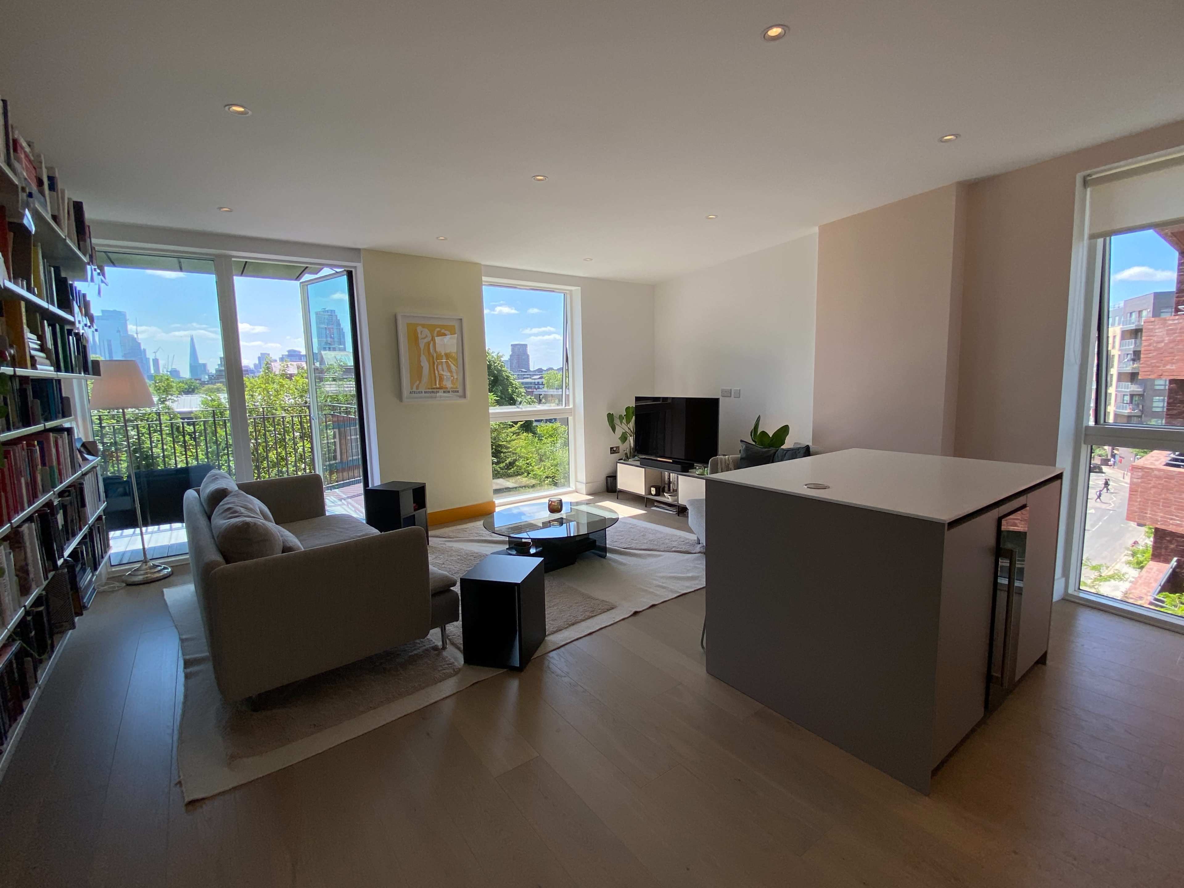 The image shows a modern living room with a couch, television, and large windows overlooking a cityscape and greenery.
