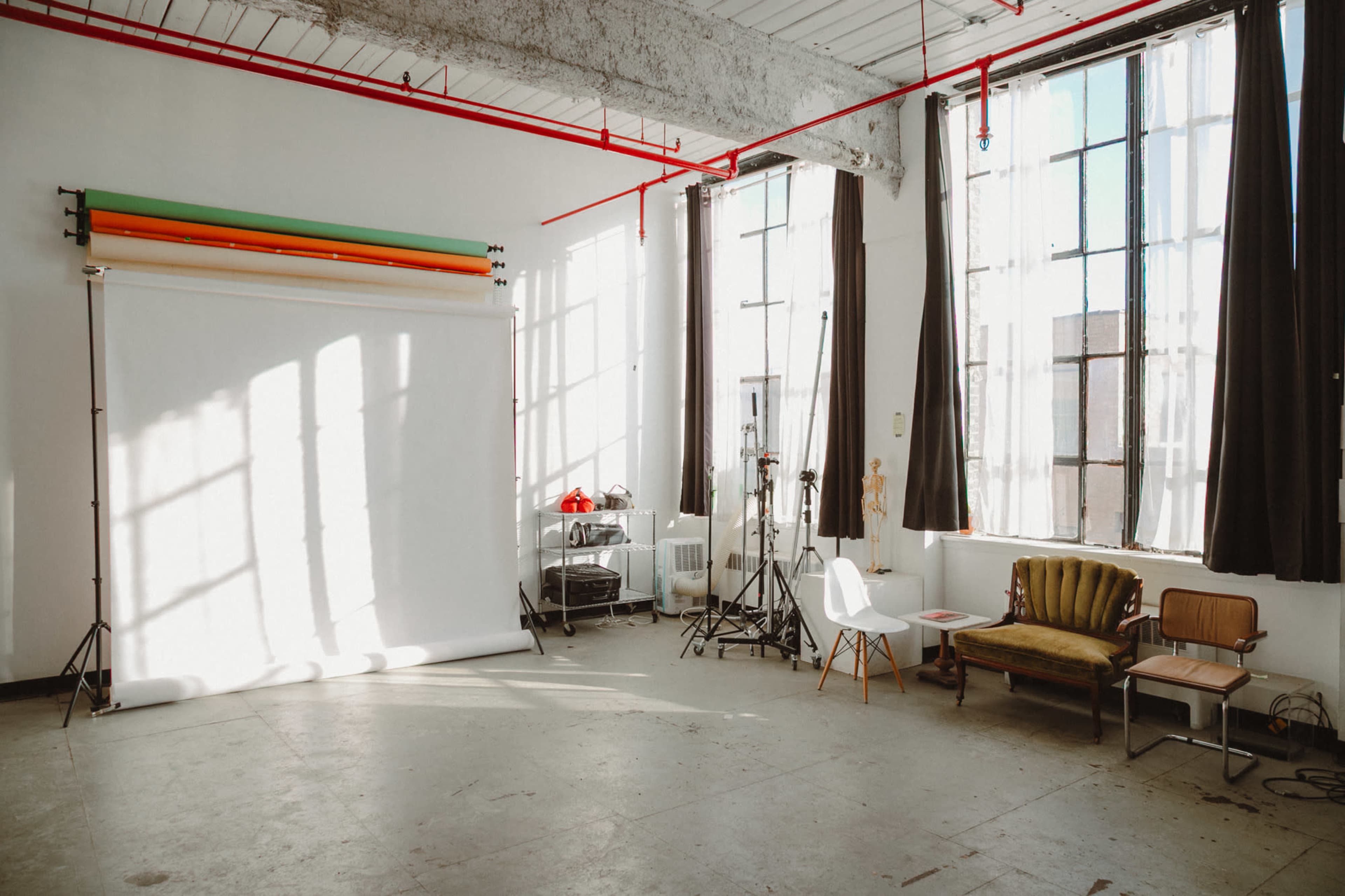 The image shows a bright photo studio with large windows, a white backdrop setup, and various furniture including a chair and a couch.