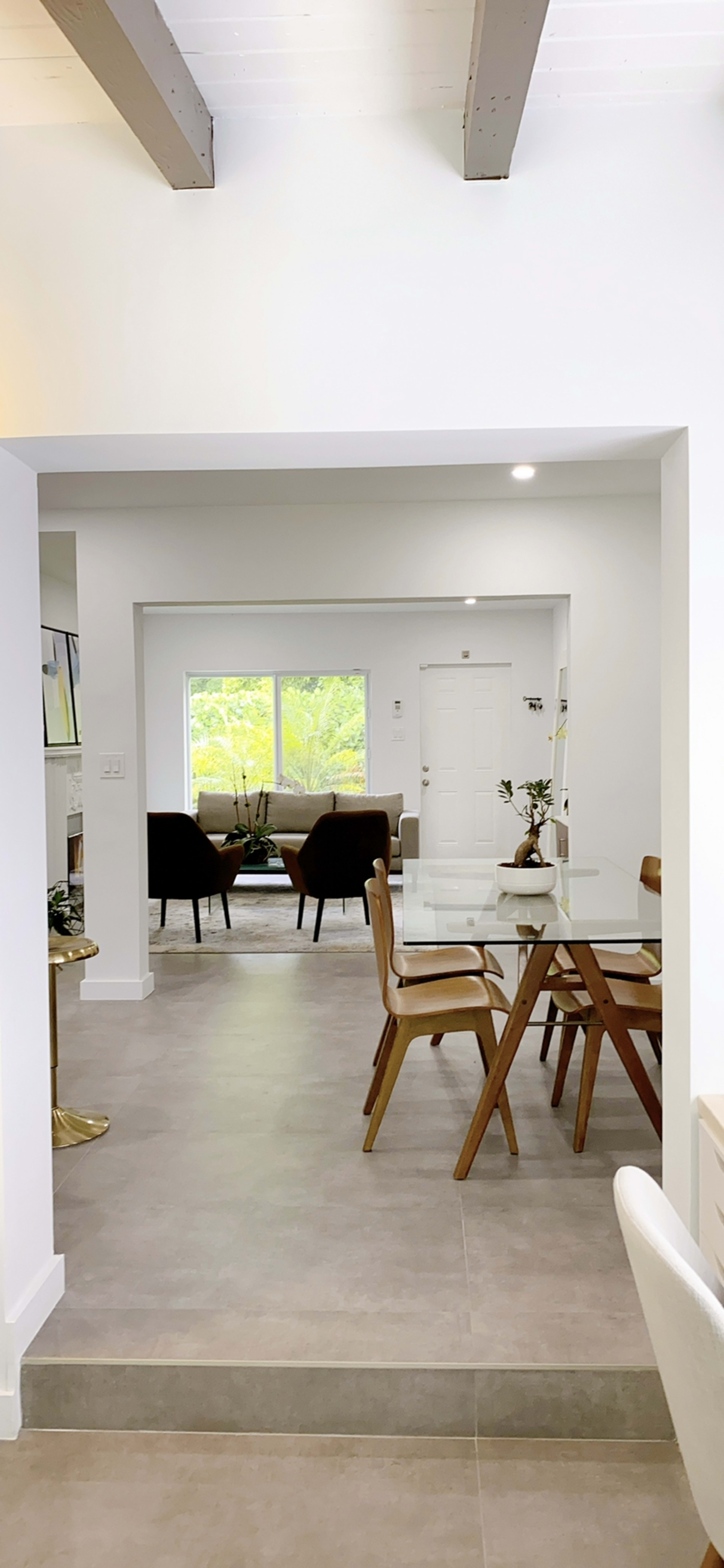 A modern interior space with a dining area featuring a table and chairs, leading to a well-lit living room with two armchairs and large windows.