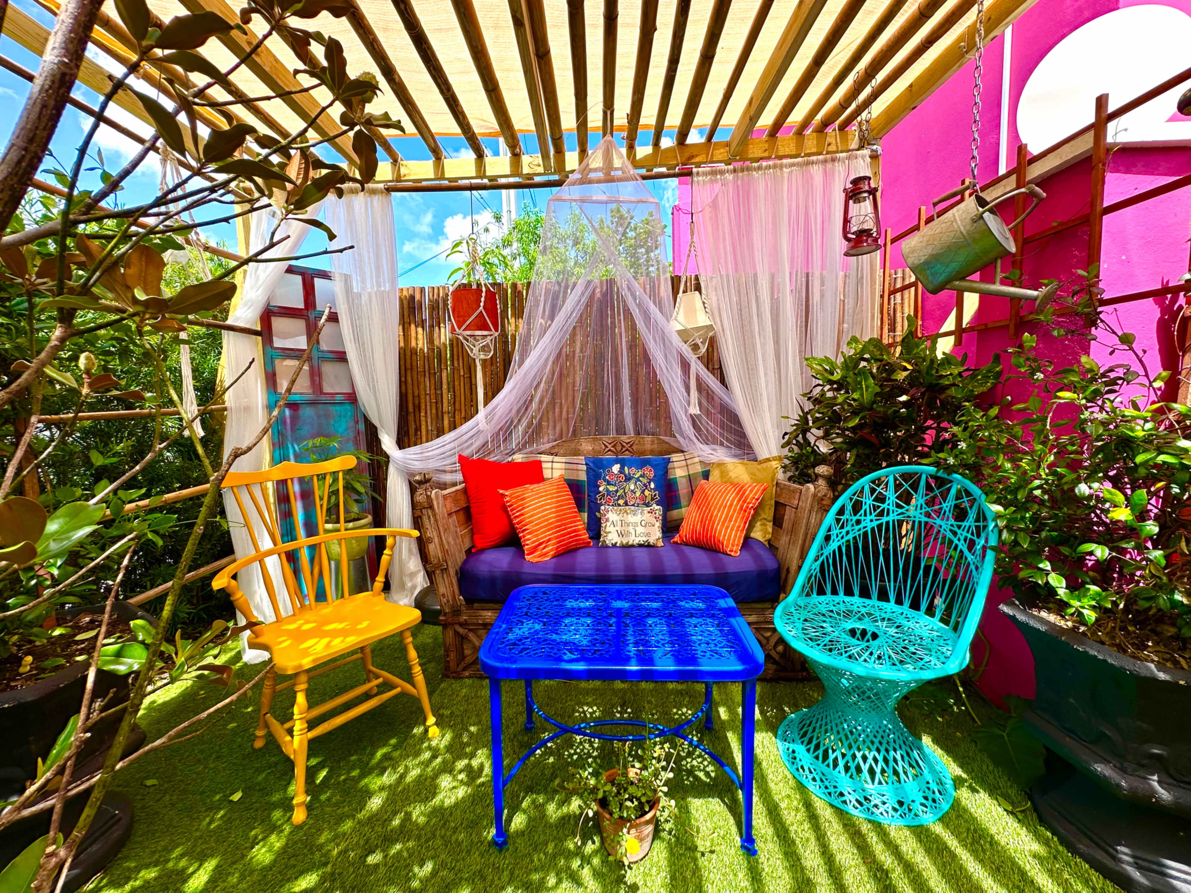 The scene features a colorful outdoor seating area with a blue table, vibrant chairs, and lush greenery under a wooden trellis.