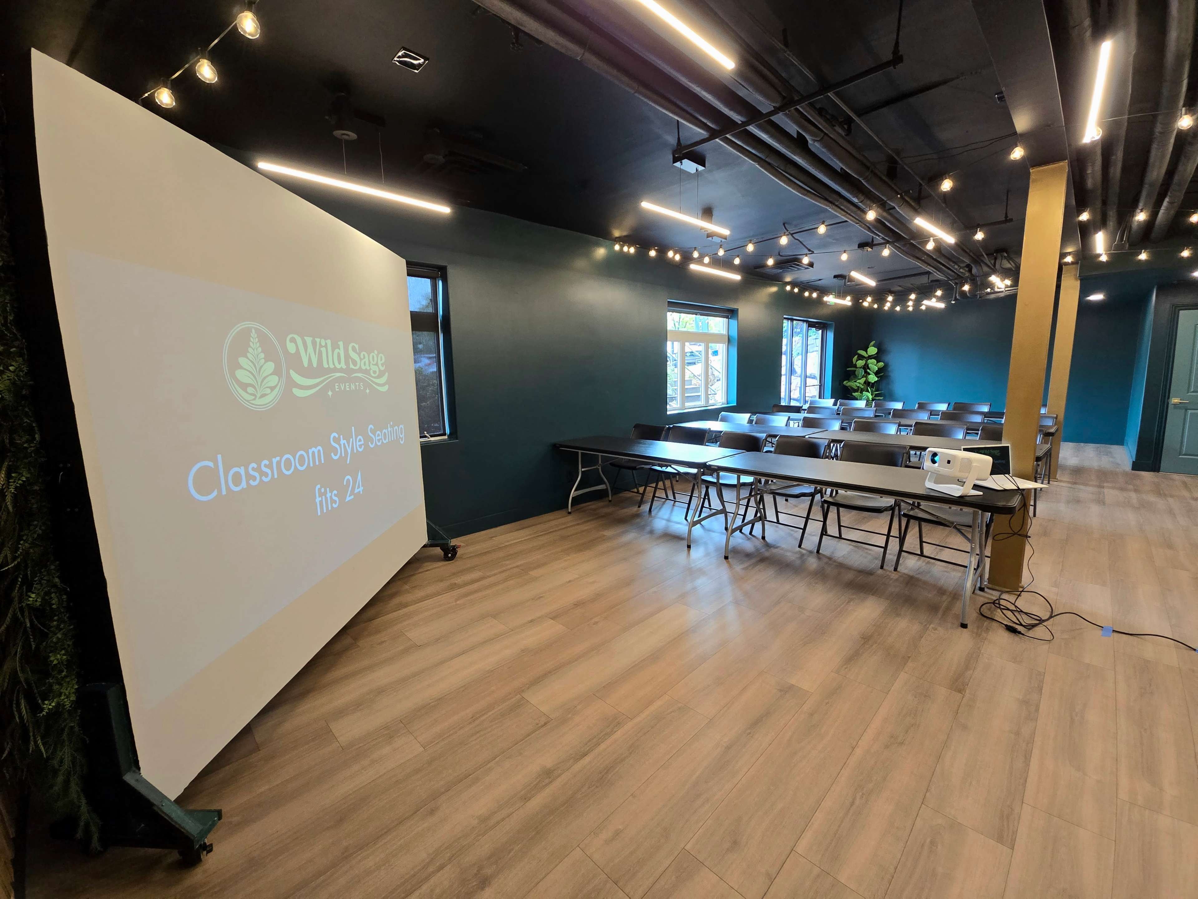 The image shows a modern classroom setting with multiple tables arranged in a classroom style and a projector displaying information on a large screen.