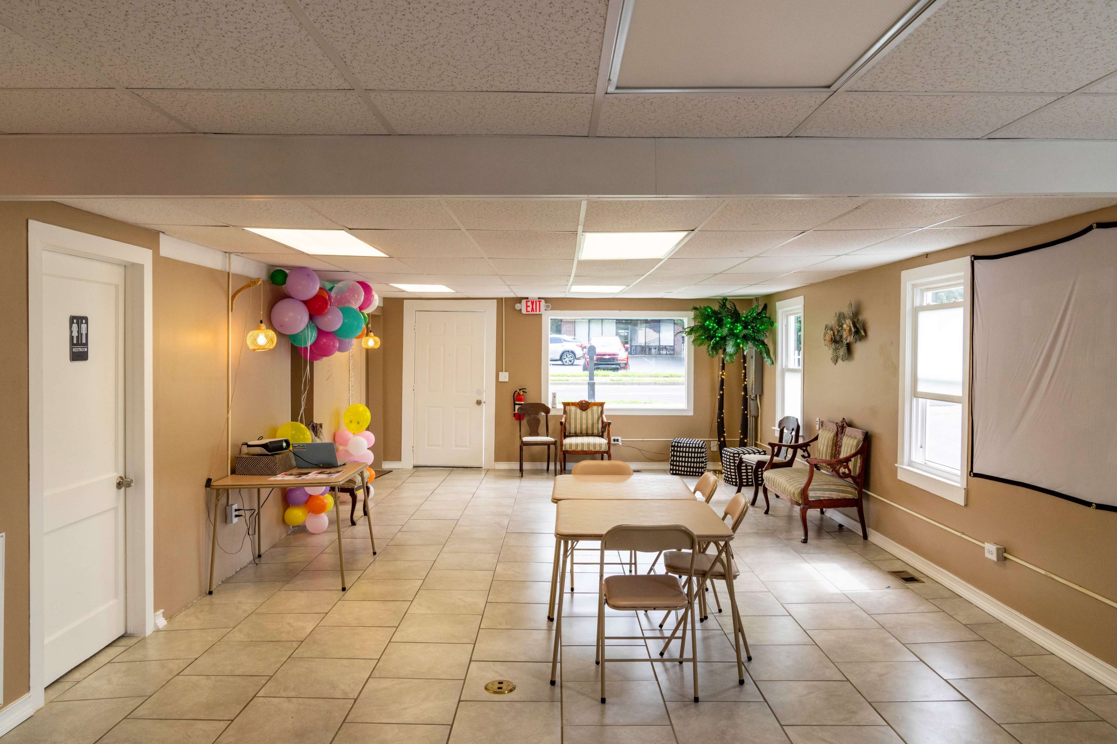 A spacious, well-lit indoor area with tables and chairs arranged, balloons attached to a wall, and a projector set up on a table.