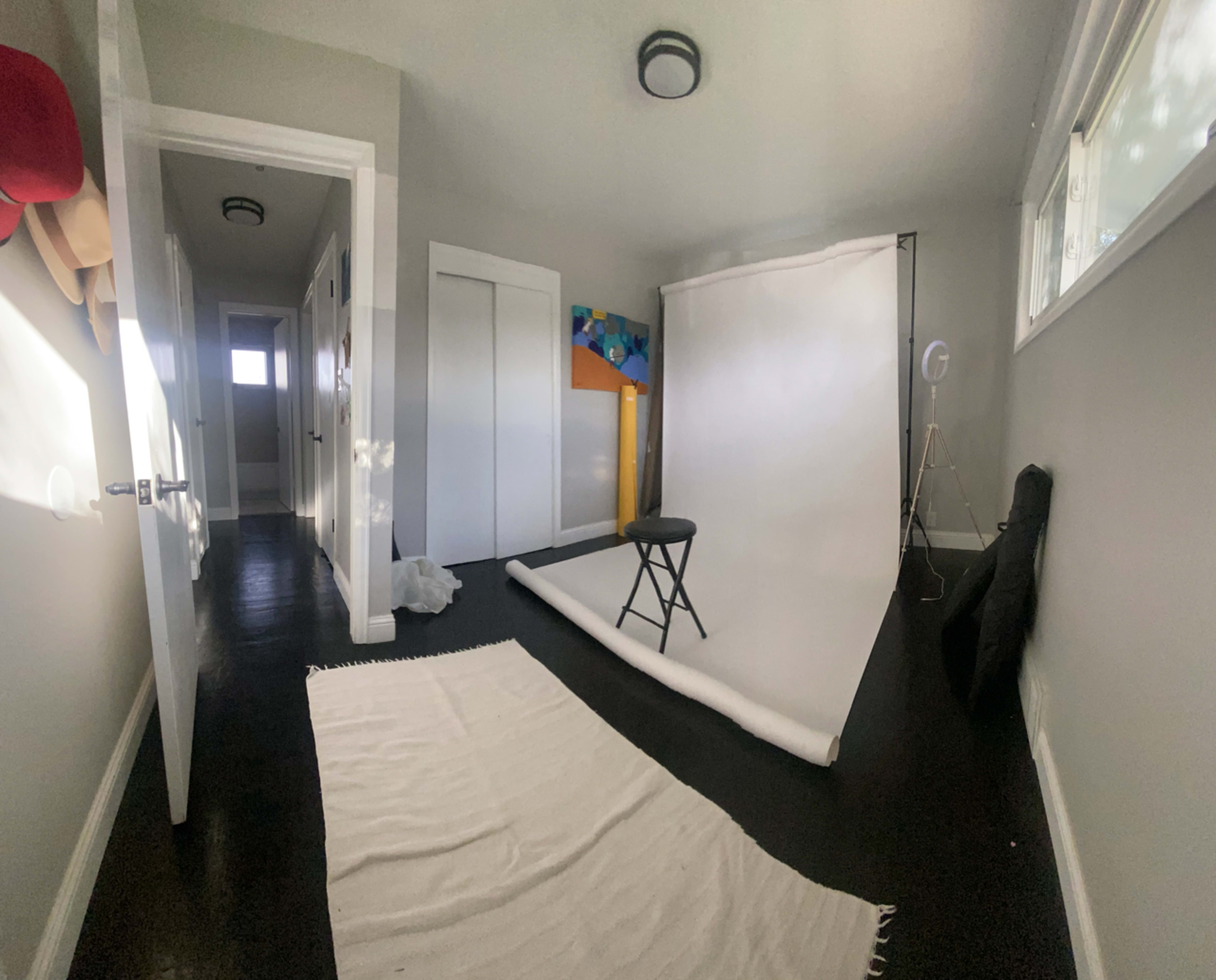 A photo studio setup with a white backdrop, a stool, and a roll of fabric on a dark floor, with a door and a hallway visible in the background.