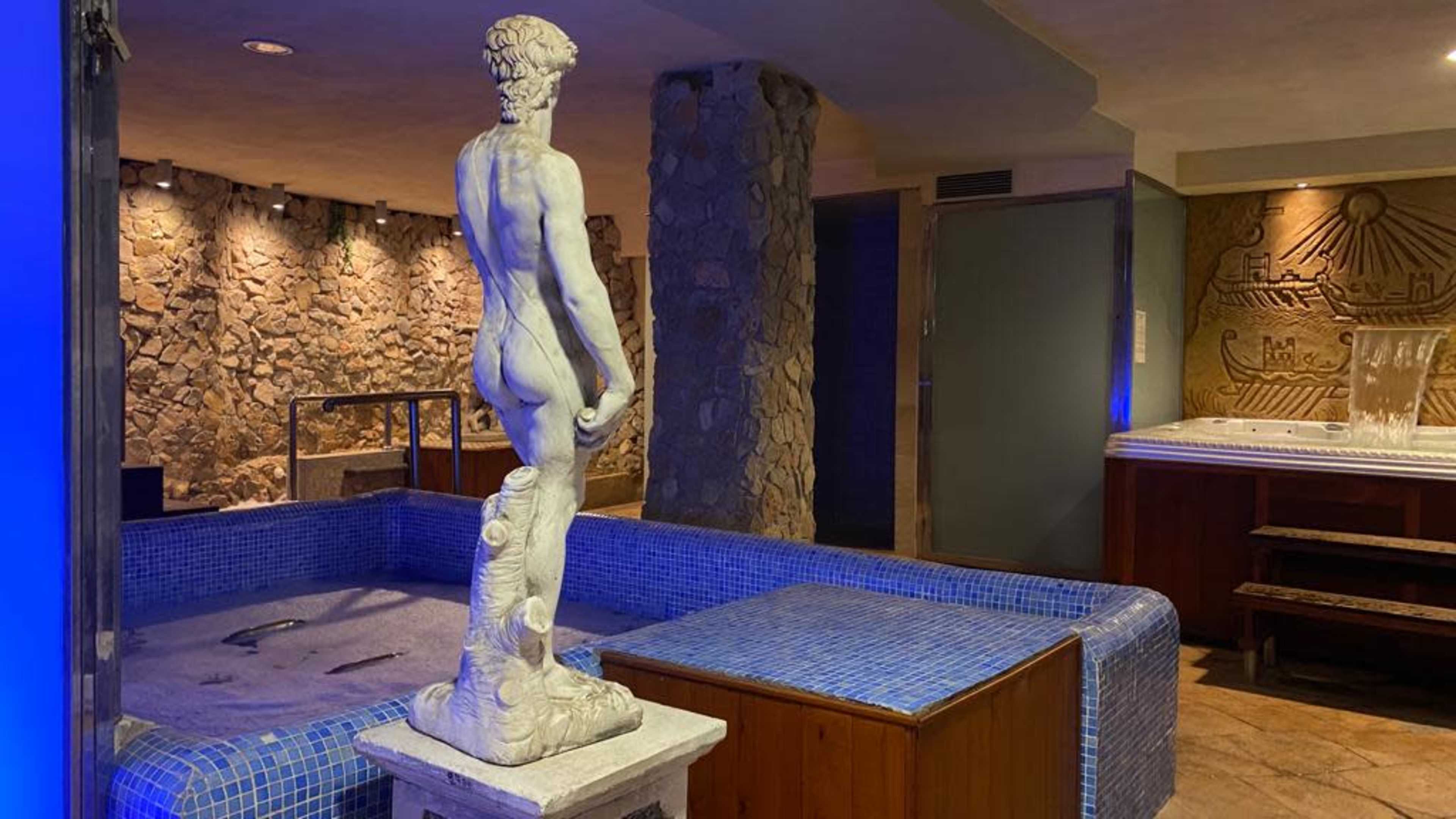 A marble statue of a nude male figure stands beside a blue-tiled hot tub in a spa-like setting with stone walls.