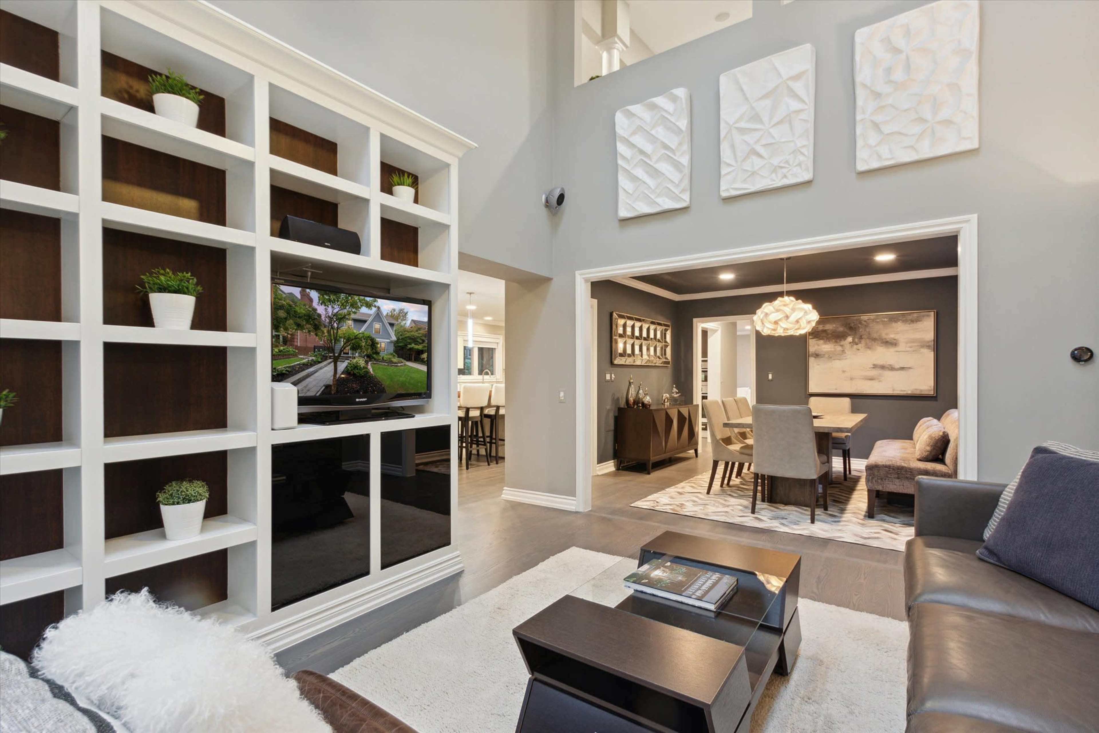 A modern living room features a built-in shelving unit with a TV, a sectional sofa, and a dining area visible in the background.