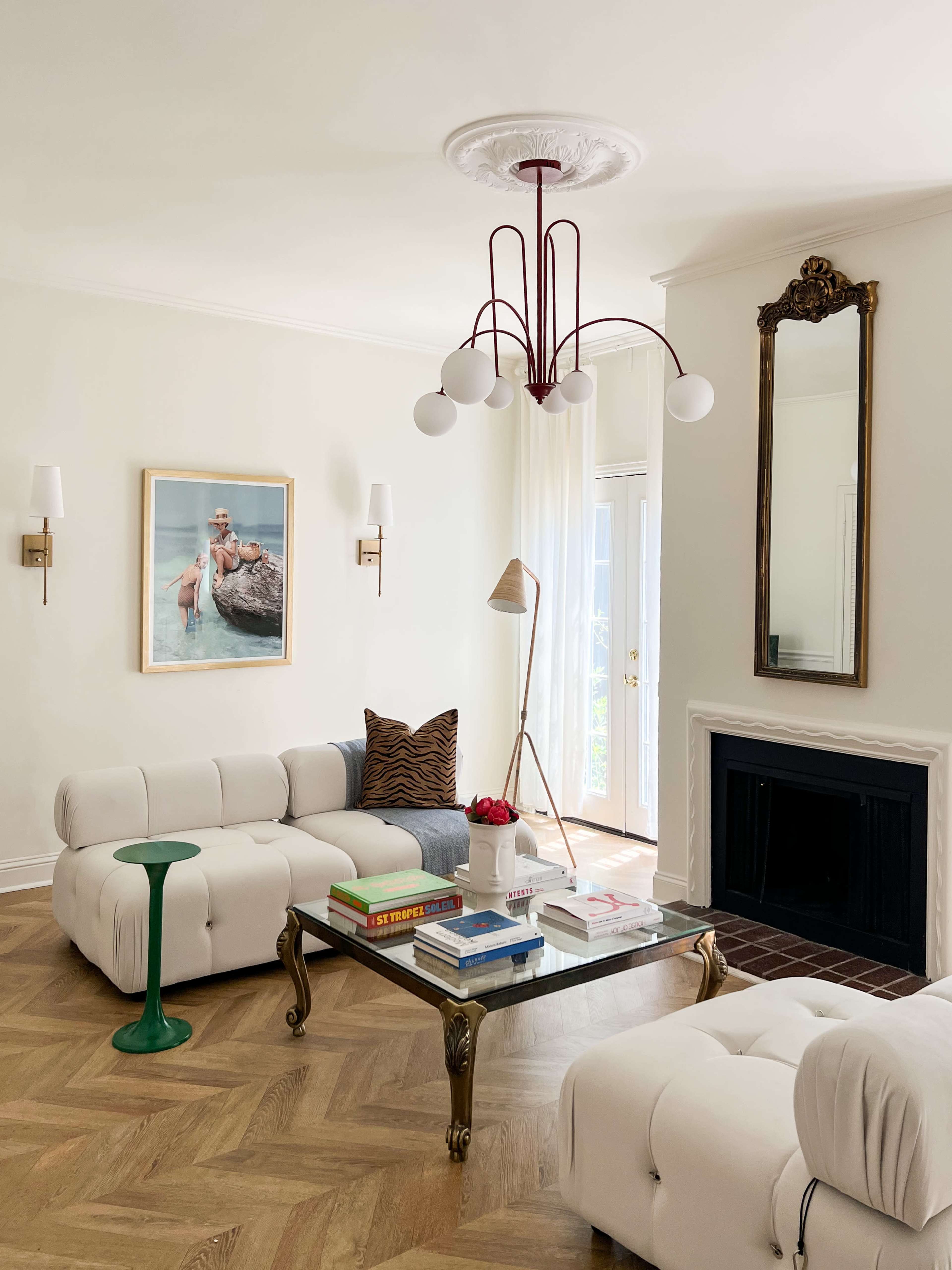 A bright living room features a white tufted sofa, a green side table, a glass coffee table topped with books, and a large ornate mirror above a fireplace.