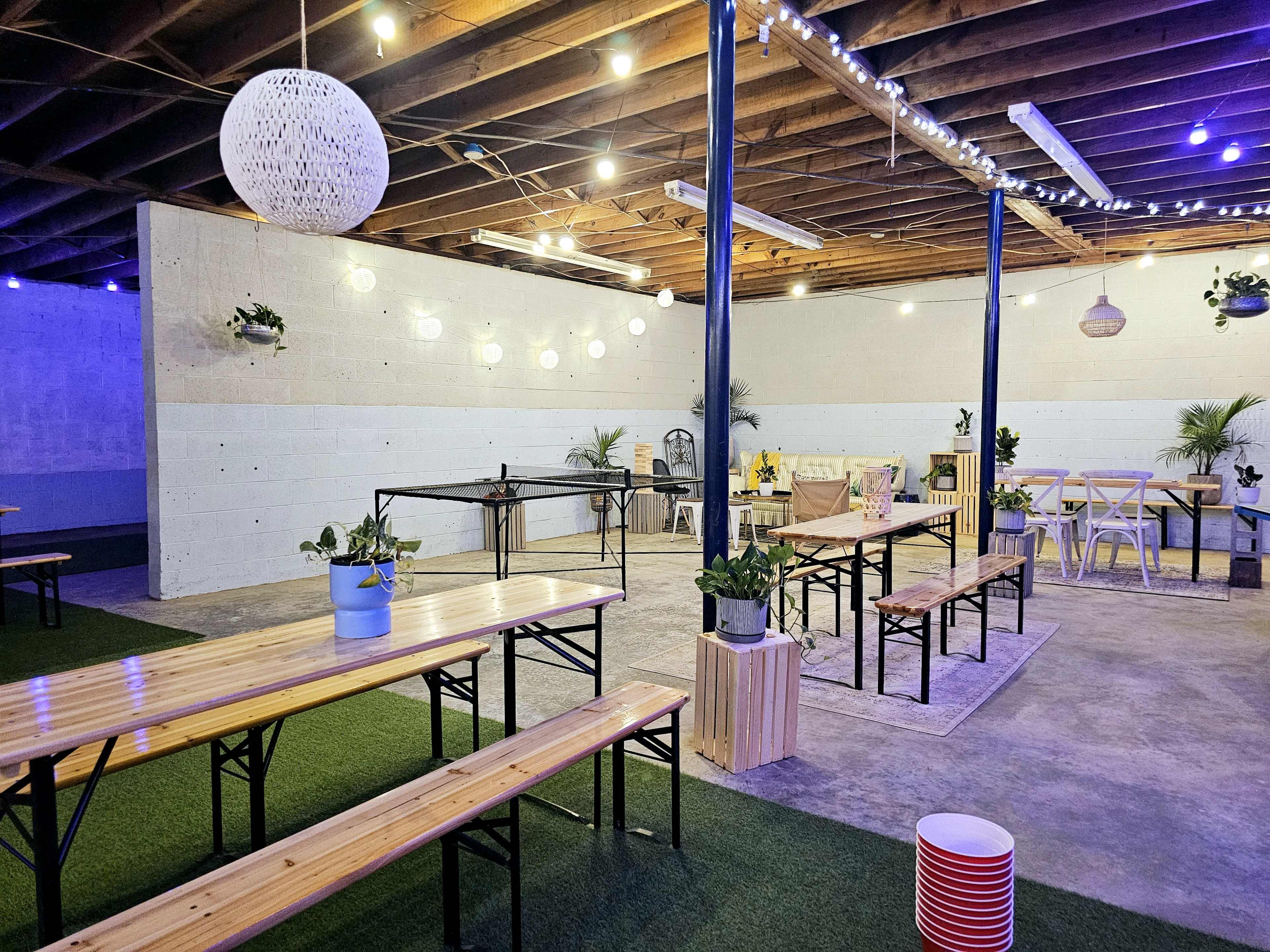 An indoor space with wooden tables and benches, green plants, and decorative lighting hanging from the ceiling.
