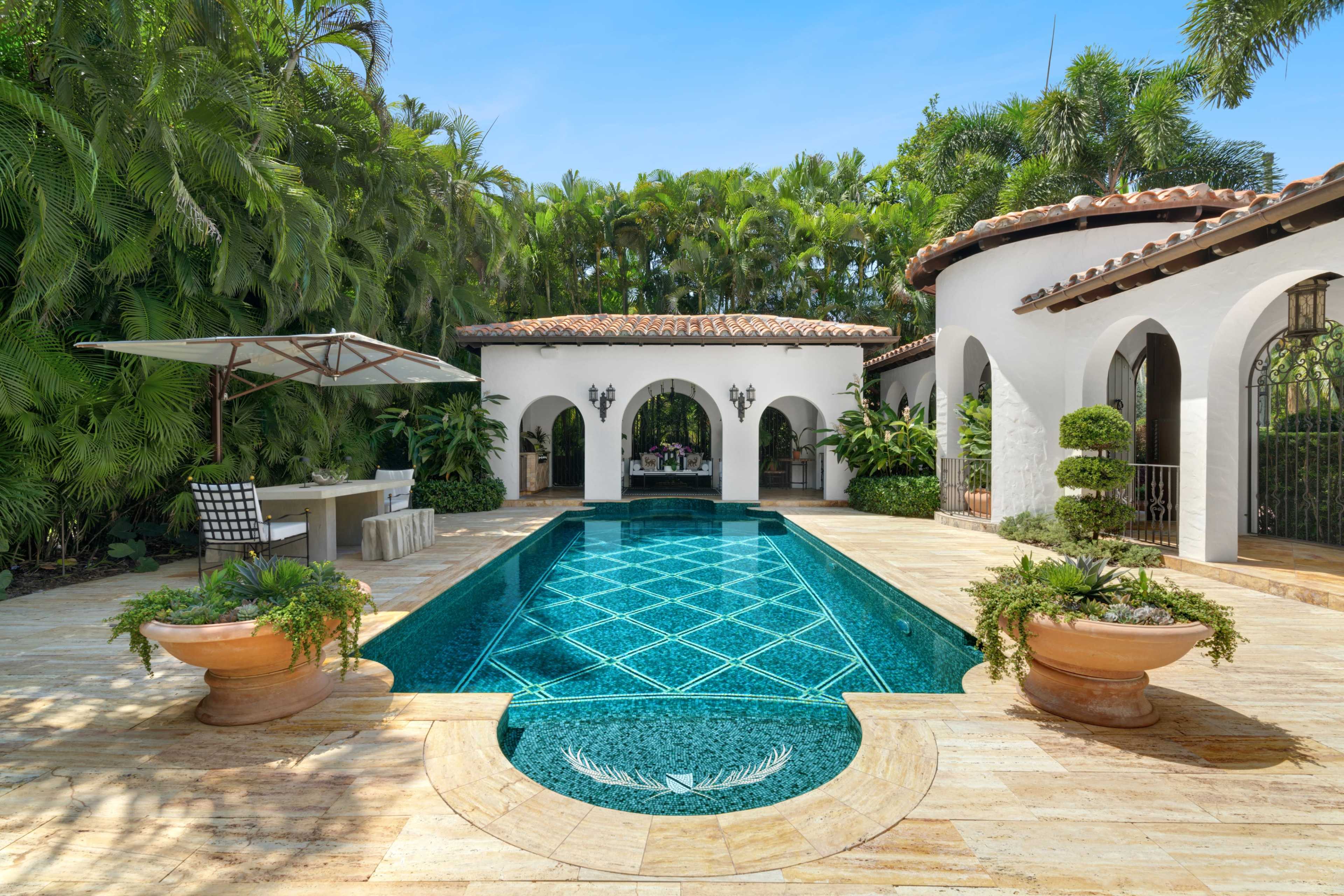 The image shows a luxurious outdoor pool area surrounded by lush greenery, featuring a tiled pool, a covered seating area, and elegant architecture.