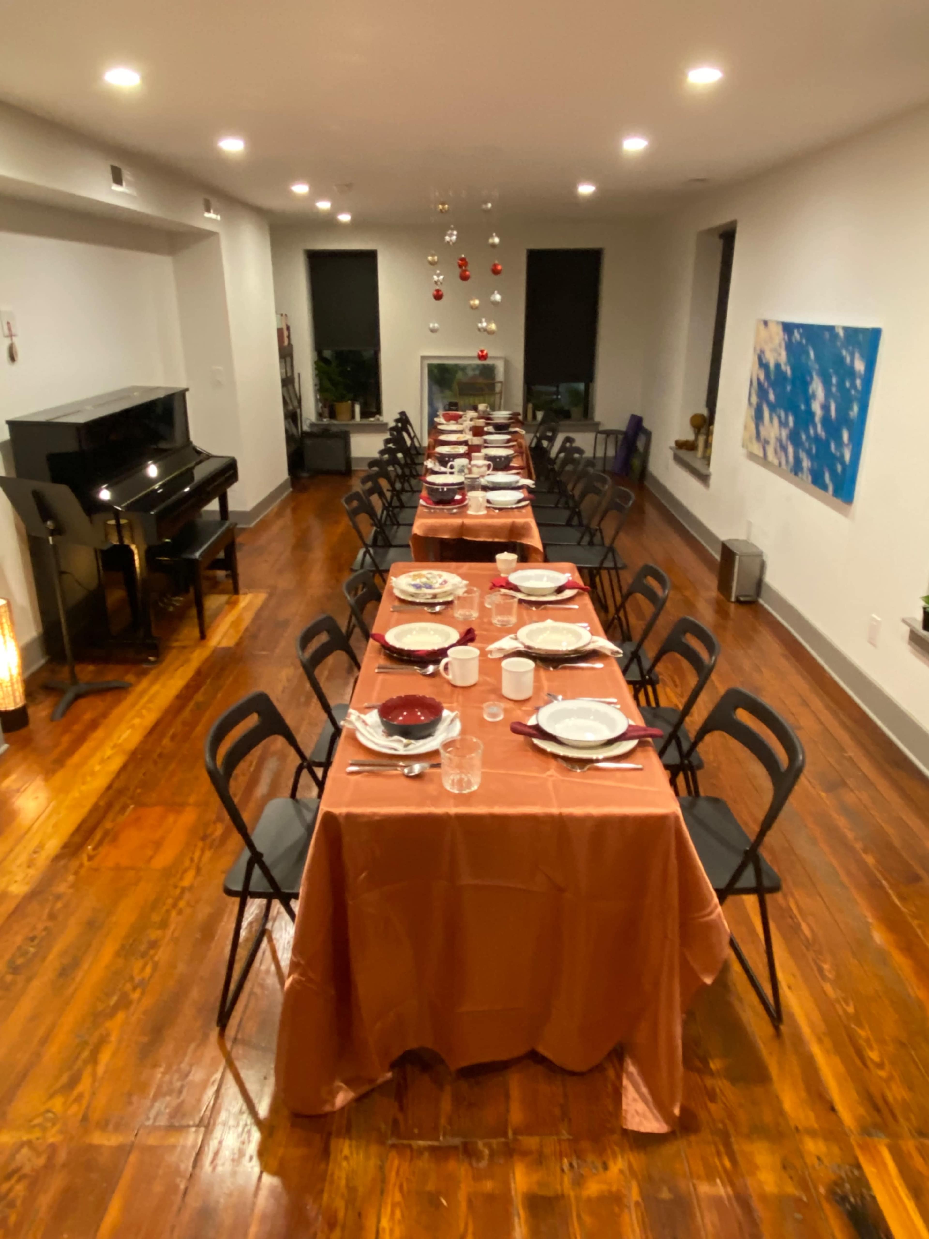 A large dining room features two long tables set with dinnerware and decorations, illuminated by overhead lights, with a piano in the corner.