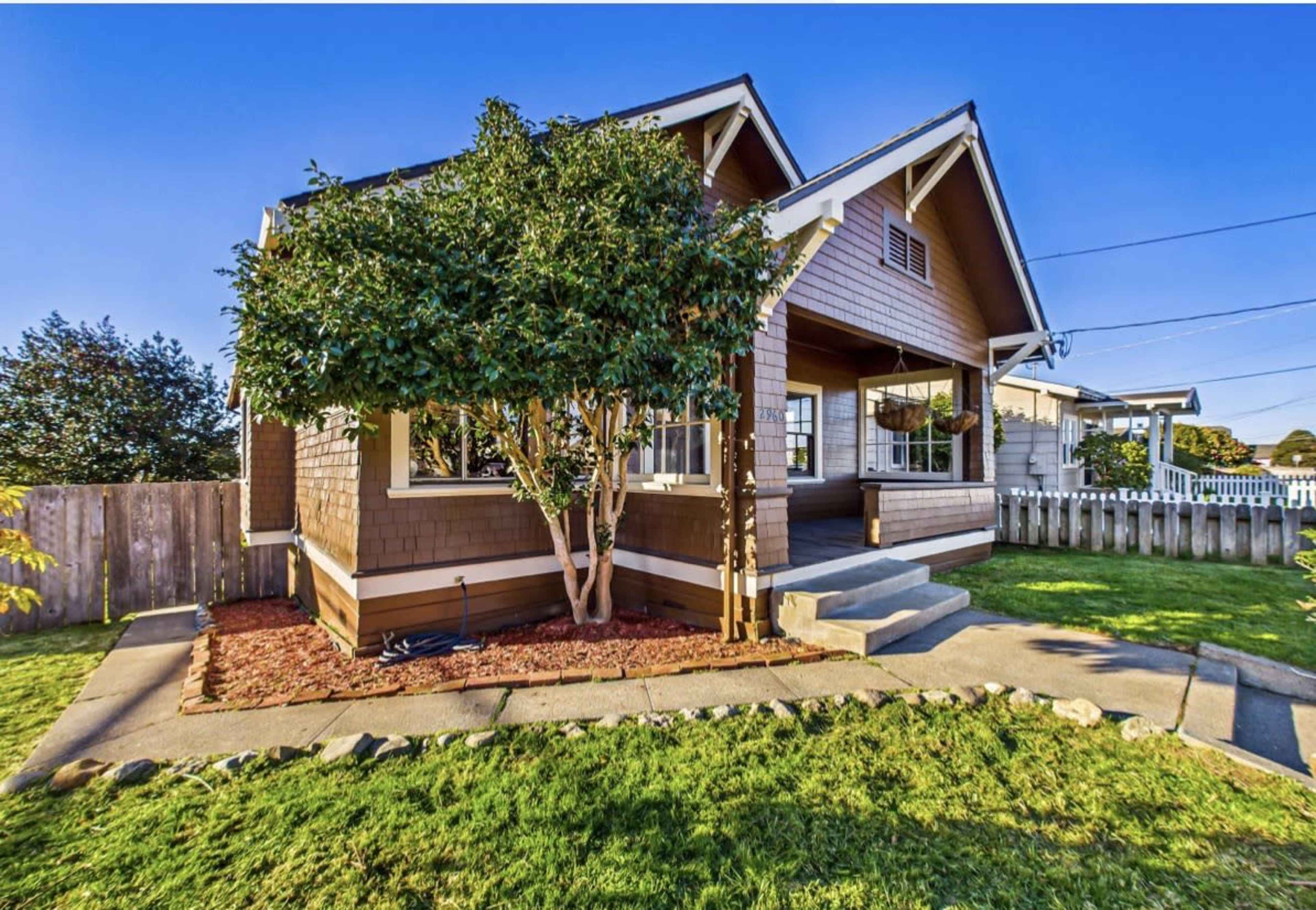 The image shows a brown house with a front porch, surrounded by green grass and a small tree in the yard.