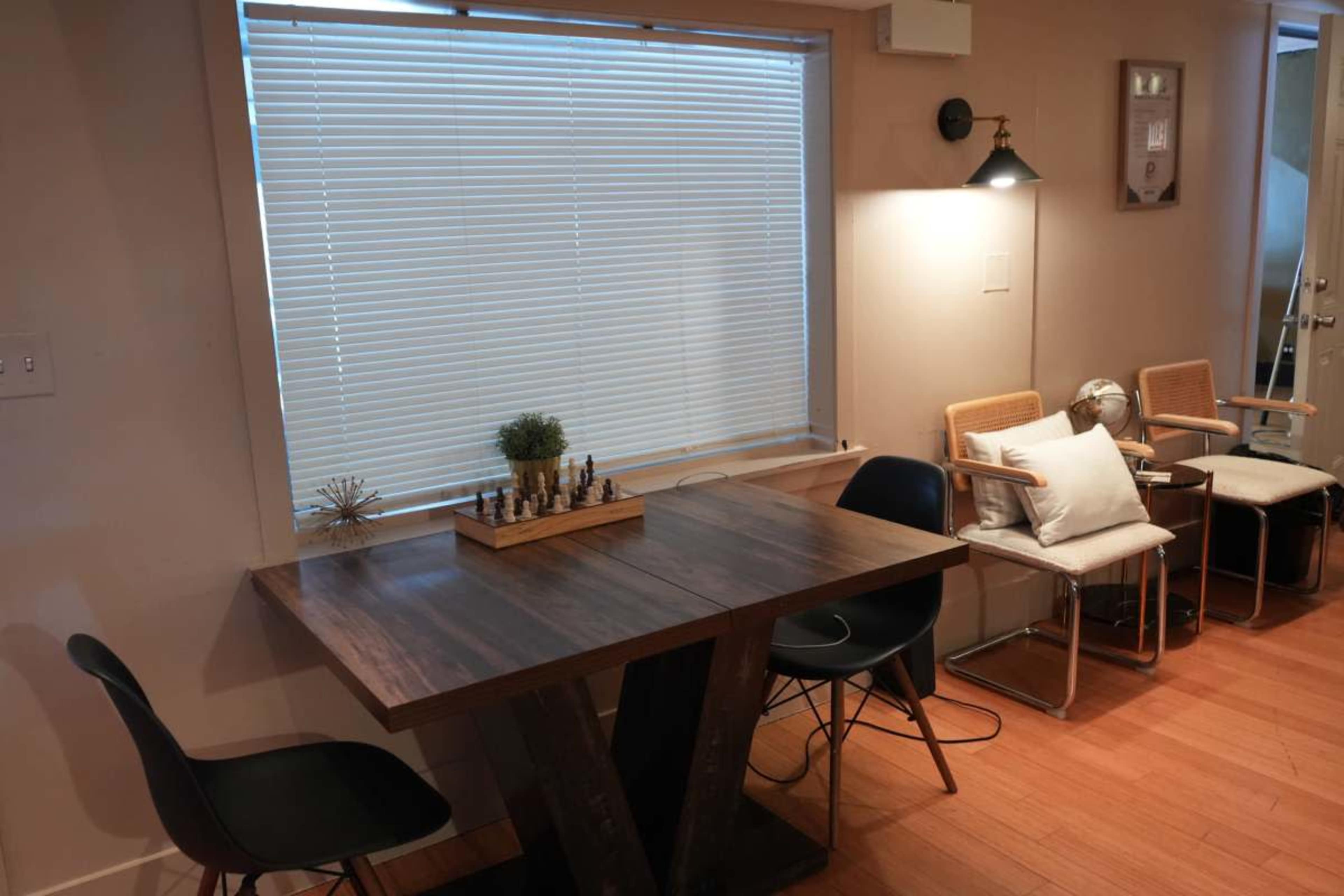 A wooden table with a chess set sits near a window with blinds, accompanied by two chairs and a small decorative plant.