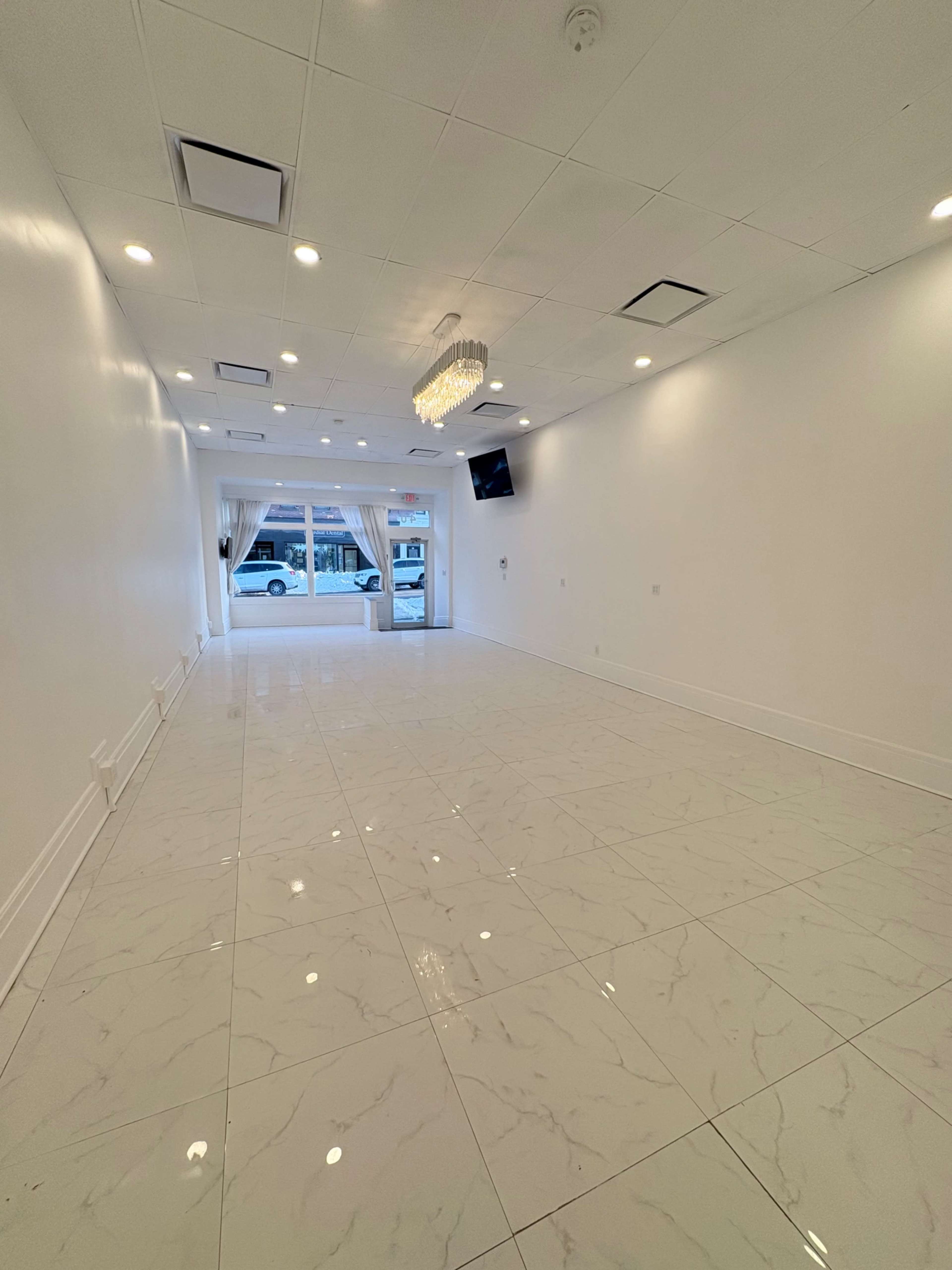 The image shows a spacious, empty room with white walls and a glossy tiled floor, featuring a large window at one end and a chandelier overhead.
