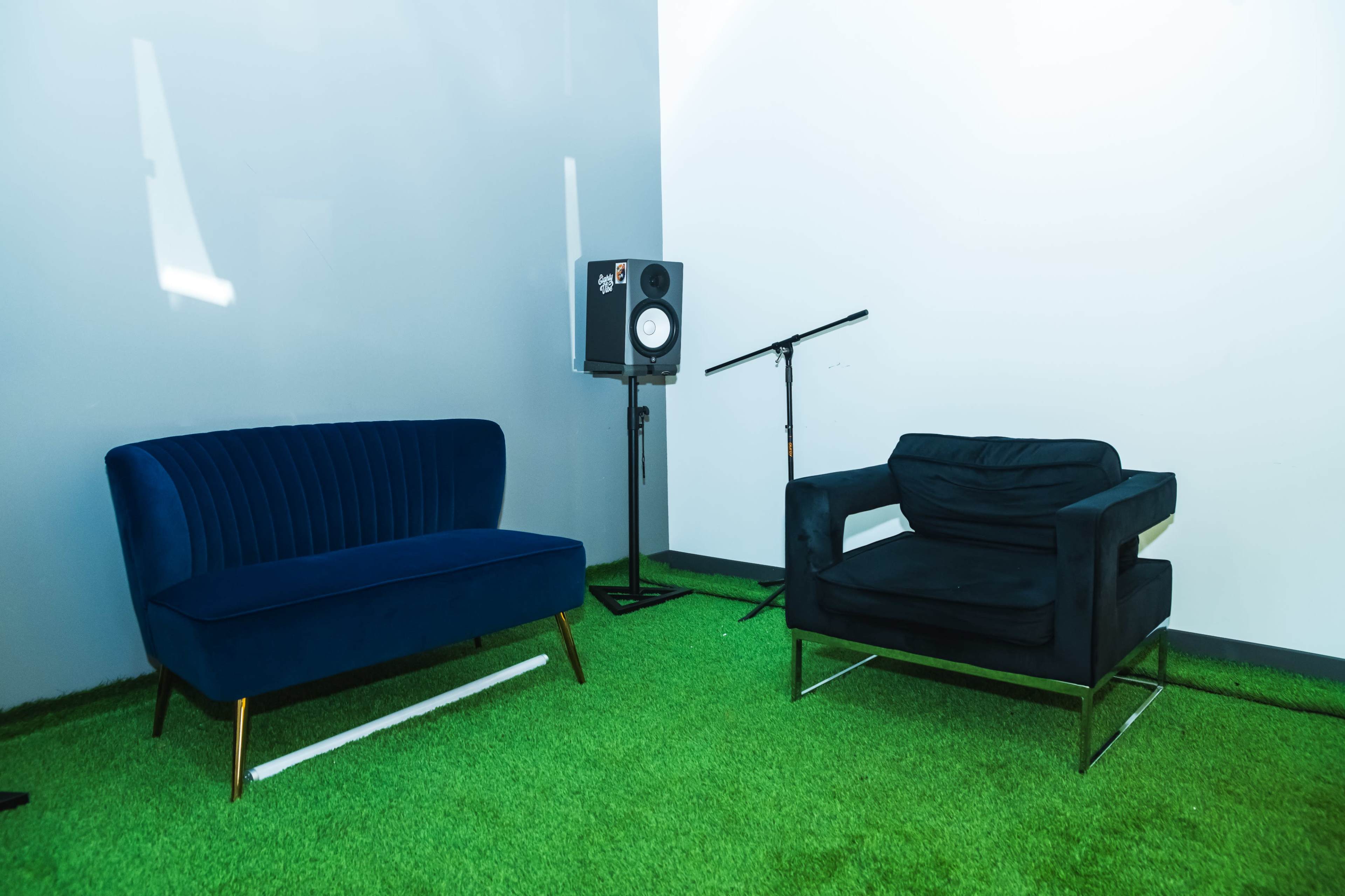 The image shows a seating area with a blue velvet sofa and a black armchair on grass-like carpeting, next to a speaker and microphone stand.