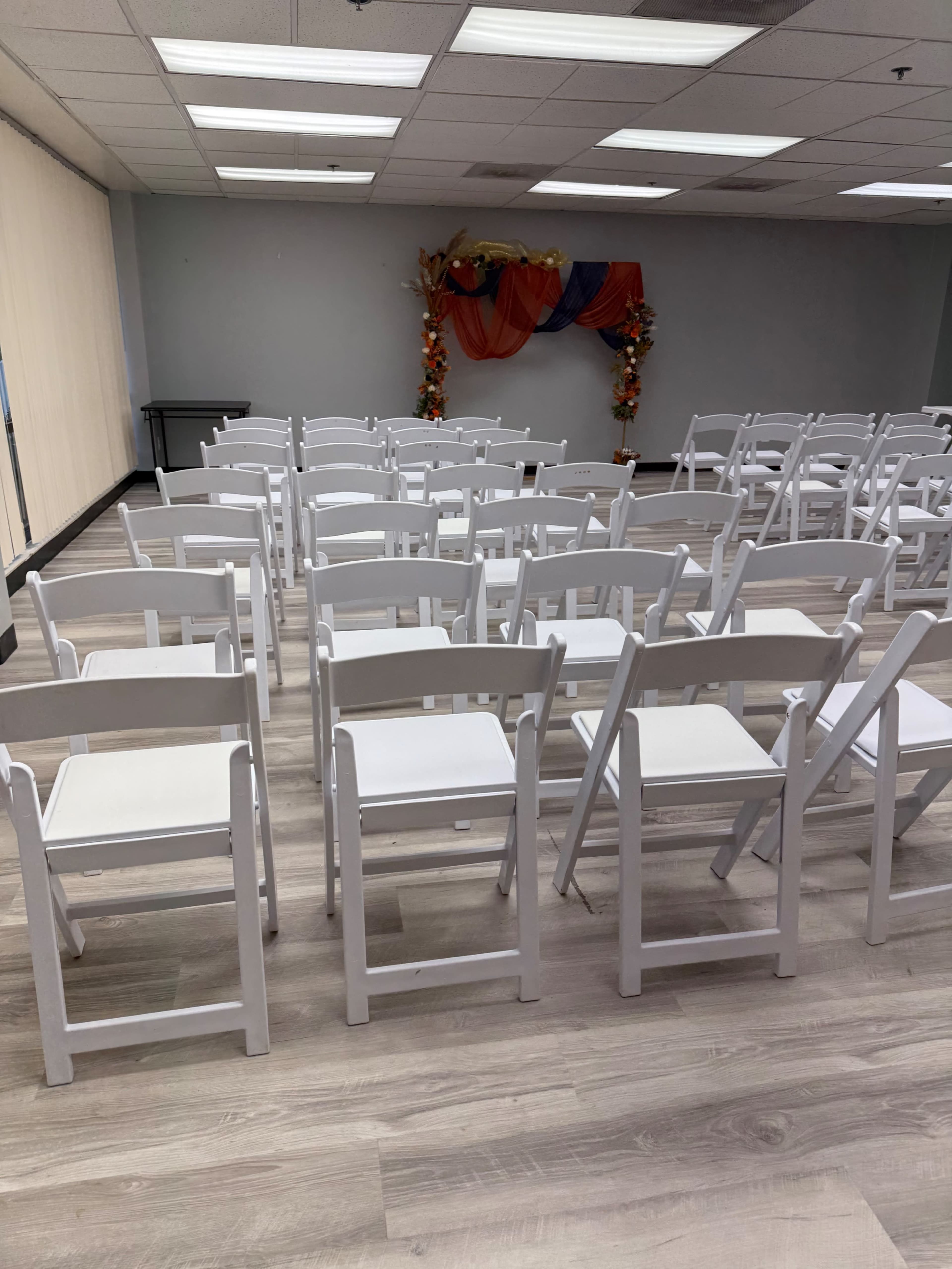 The image shows a room with rows of white folding chairs arranged for an event, with decorative fabric and foliage in the background.