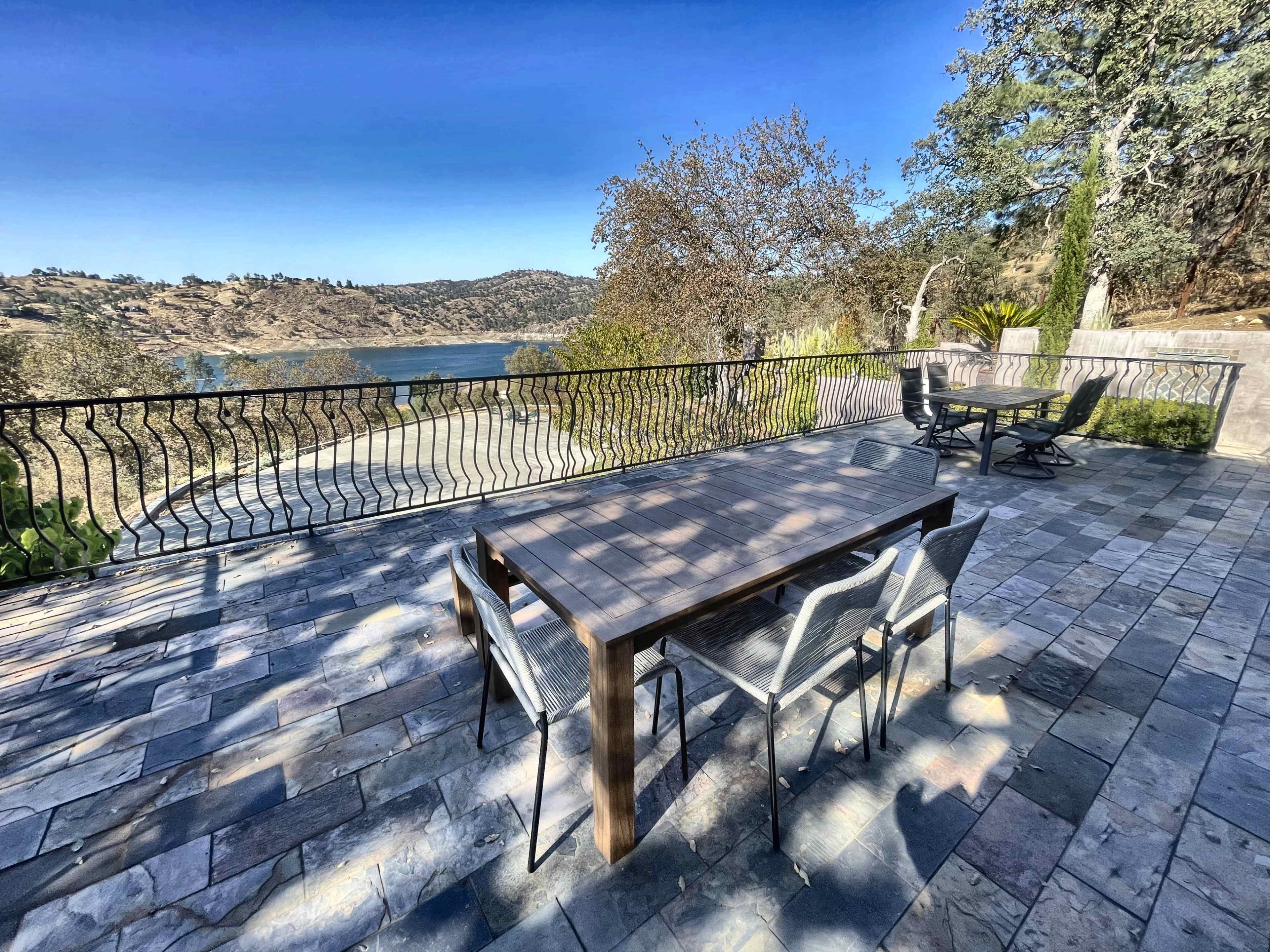 A patio area features a wooden table and chairs with a view of a lake surrounded by hills and trees.