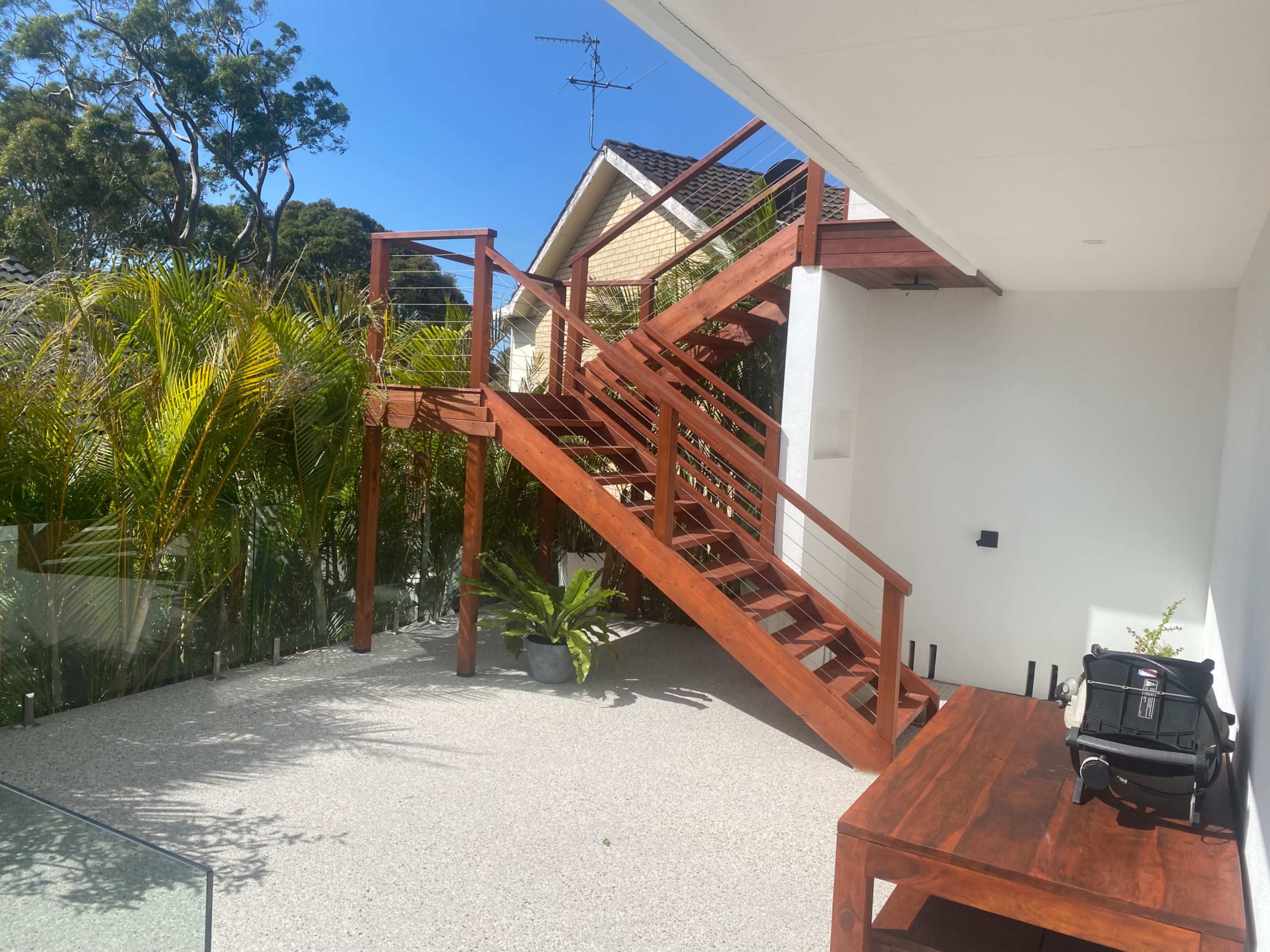 A wooden staircase with railings leads from a patio area to a higher level of a house, surrounded by greenery and featuring a table on the patio.