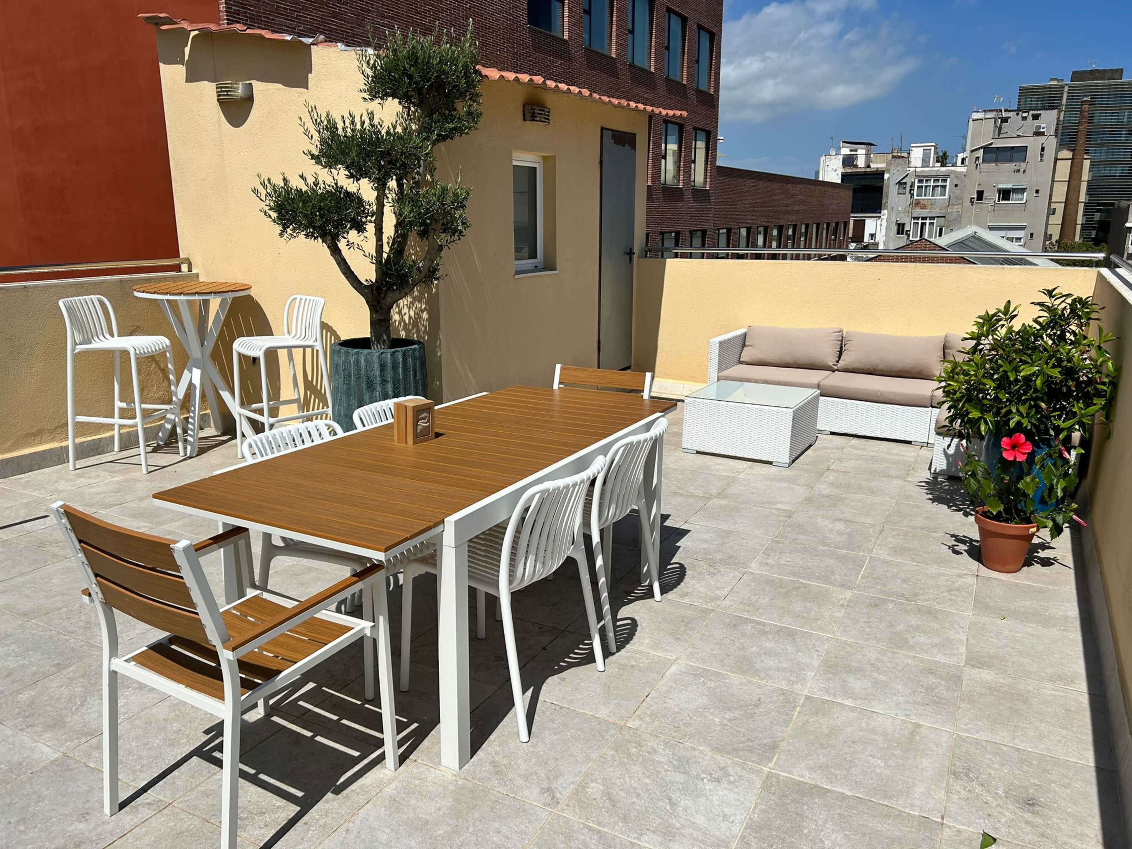 A rooftop terrace featuring a wooden table with several chairs, a couch, potted plants, and a view of surrounding buildings under a clear sky.