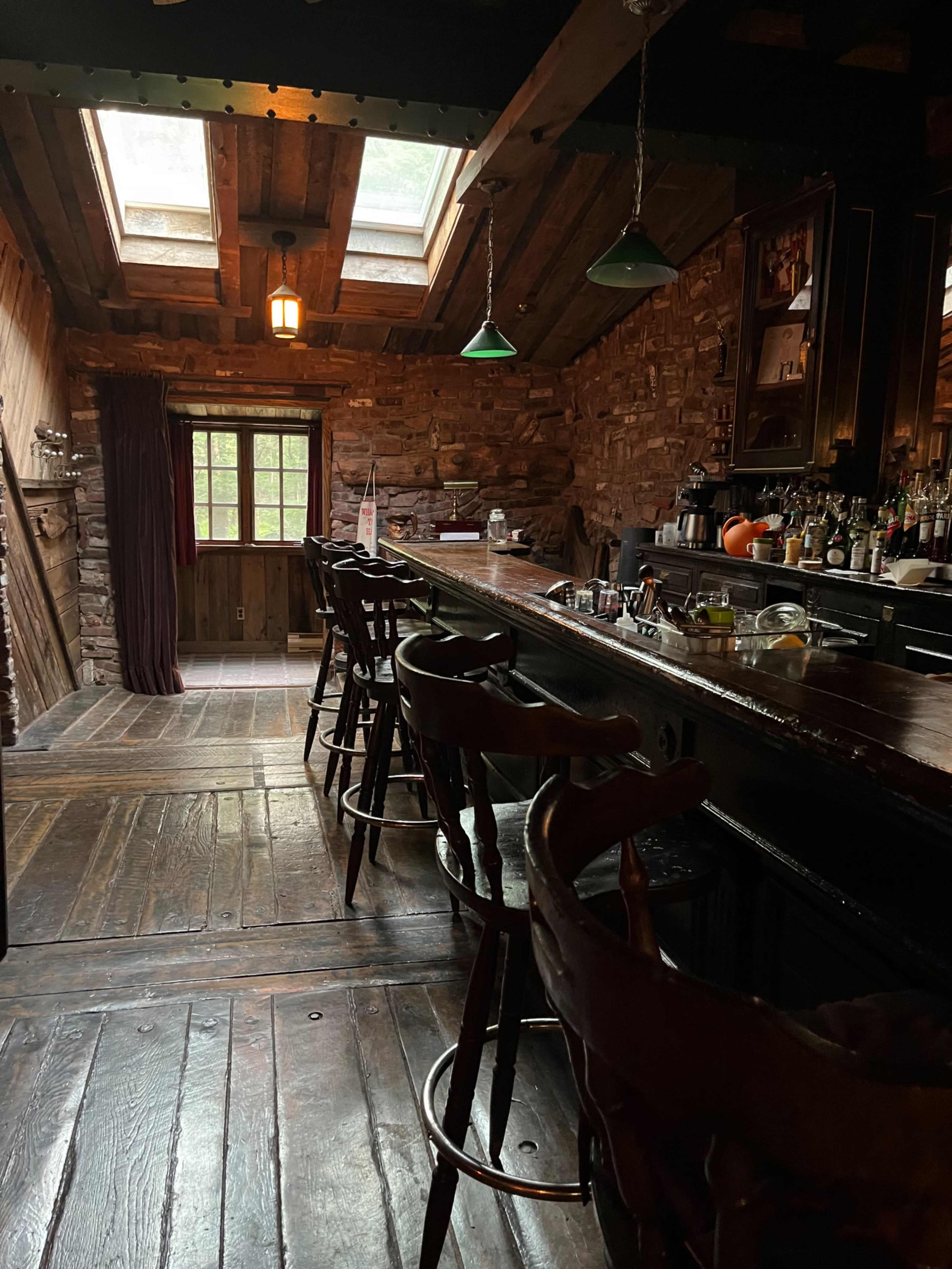 A rustic bar area with a wooden countertop, high stools, and a stone wall, illuminated by natural light from skylights above.