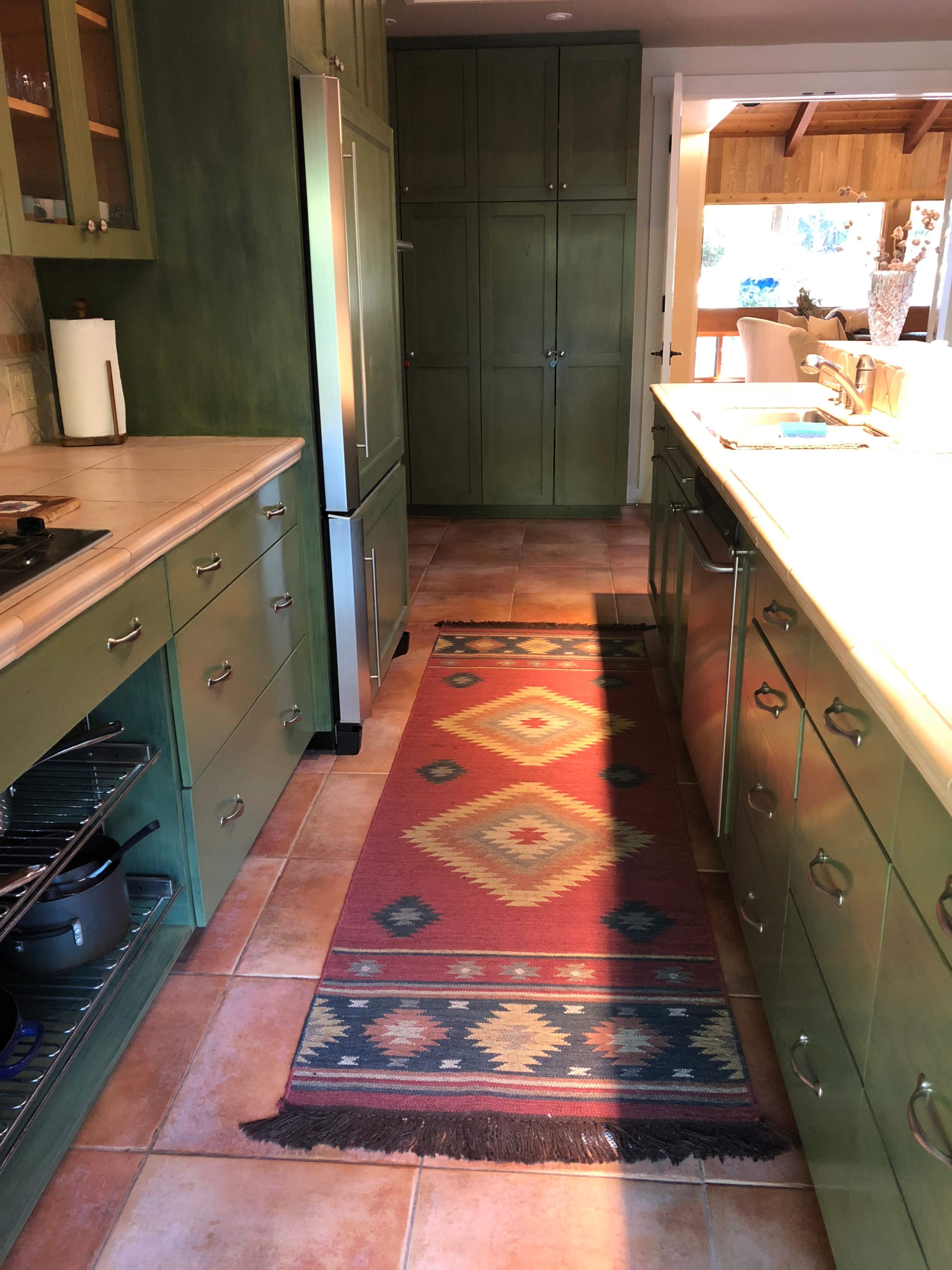 A kitchen with green cabinets, a patterned rug running the length of the floor, and an open layout leading to a dining area.