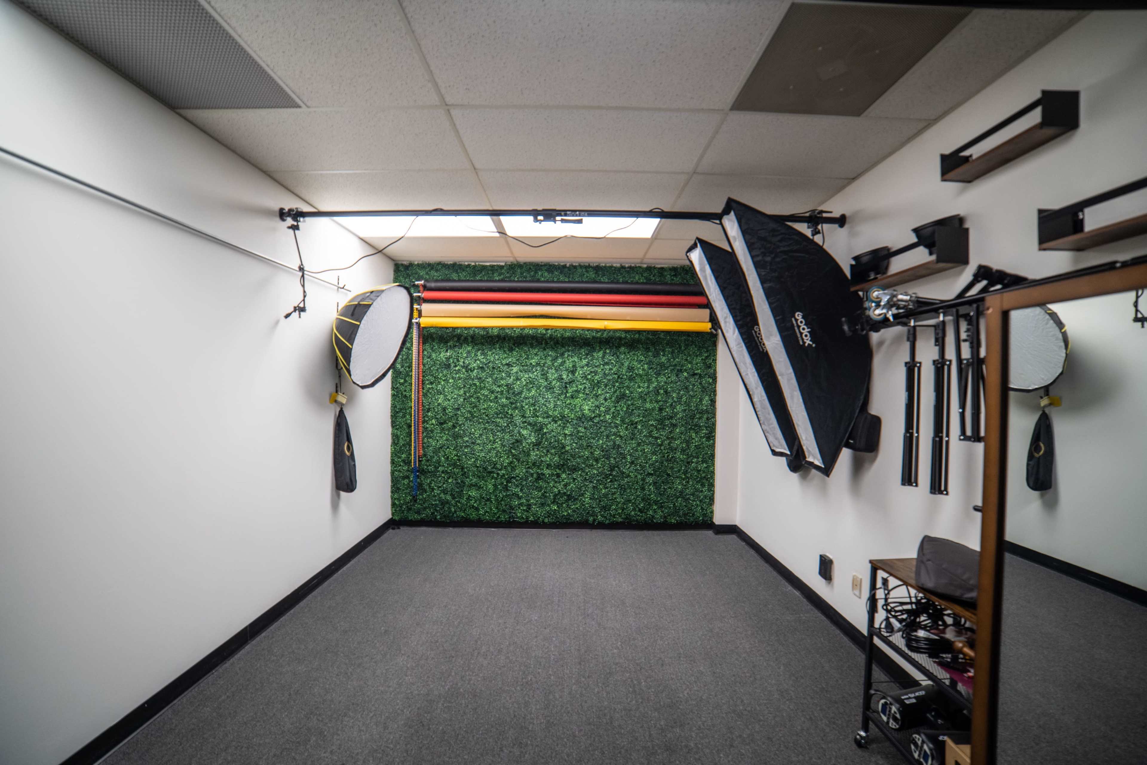 A photography studio with a green foliage backdrop, lighting equipment, and various props arranged along the walls.