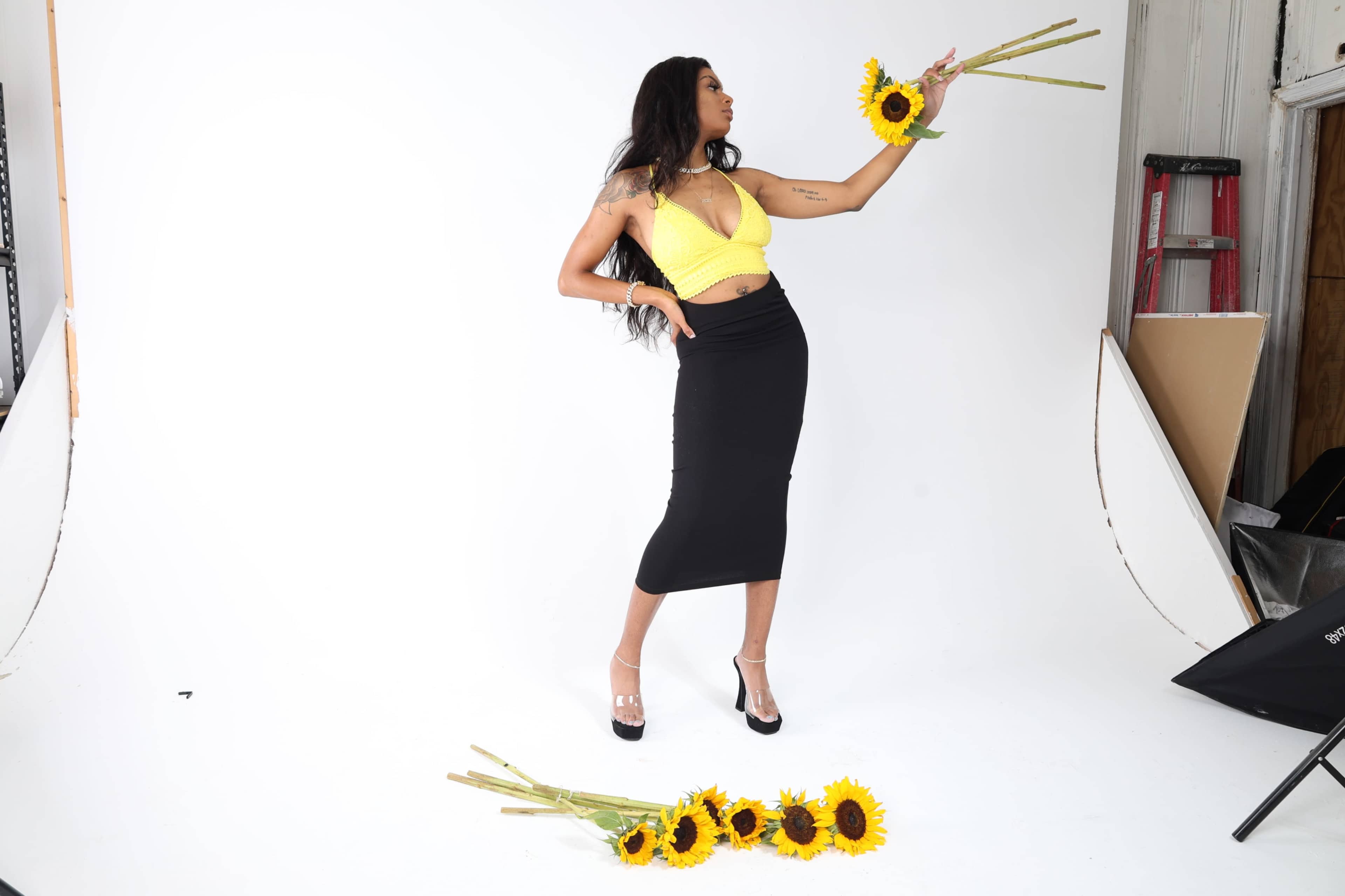 A woman in a yellow top and black skirt poses with sunflowers in a studio setting.