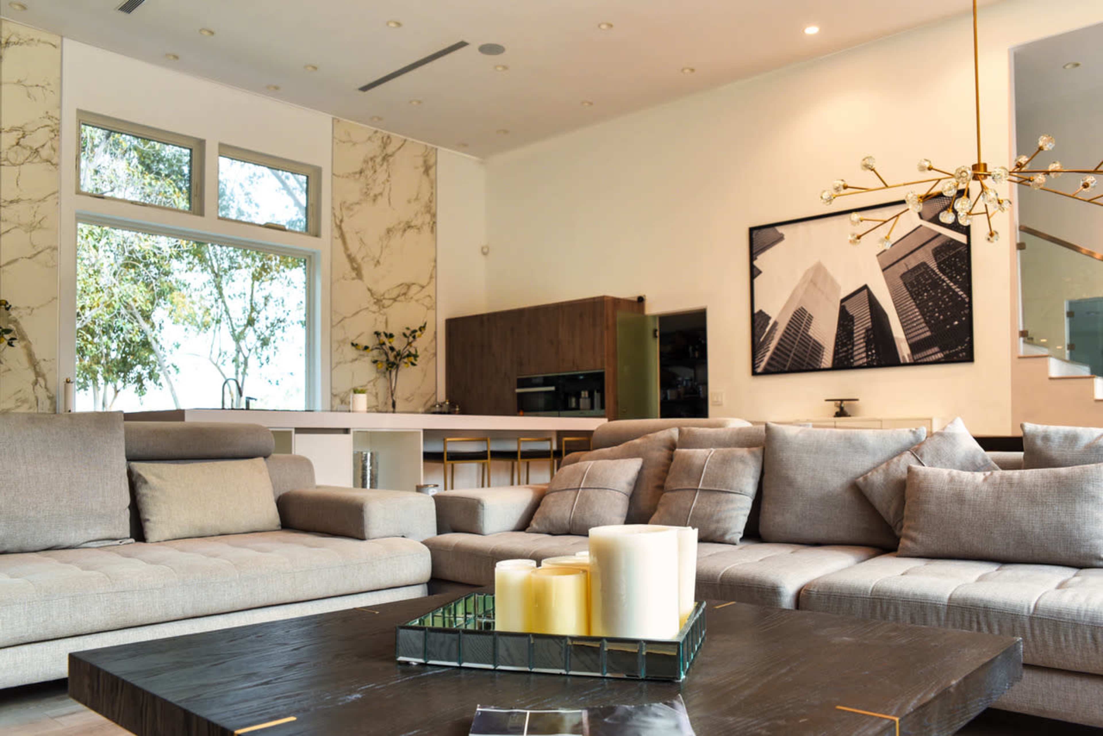 A modern living room with beige sofas, a dark wooden coffee table holding candles, and large windows that provide natural light.