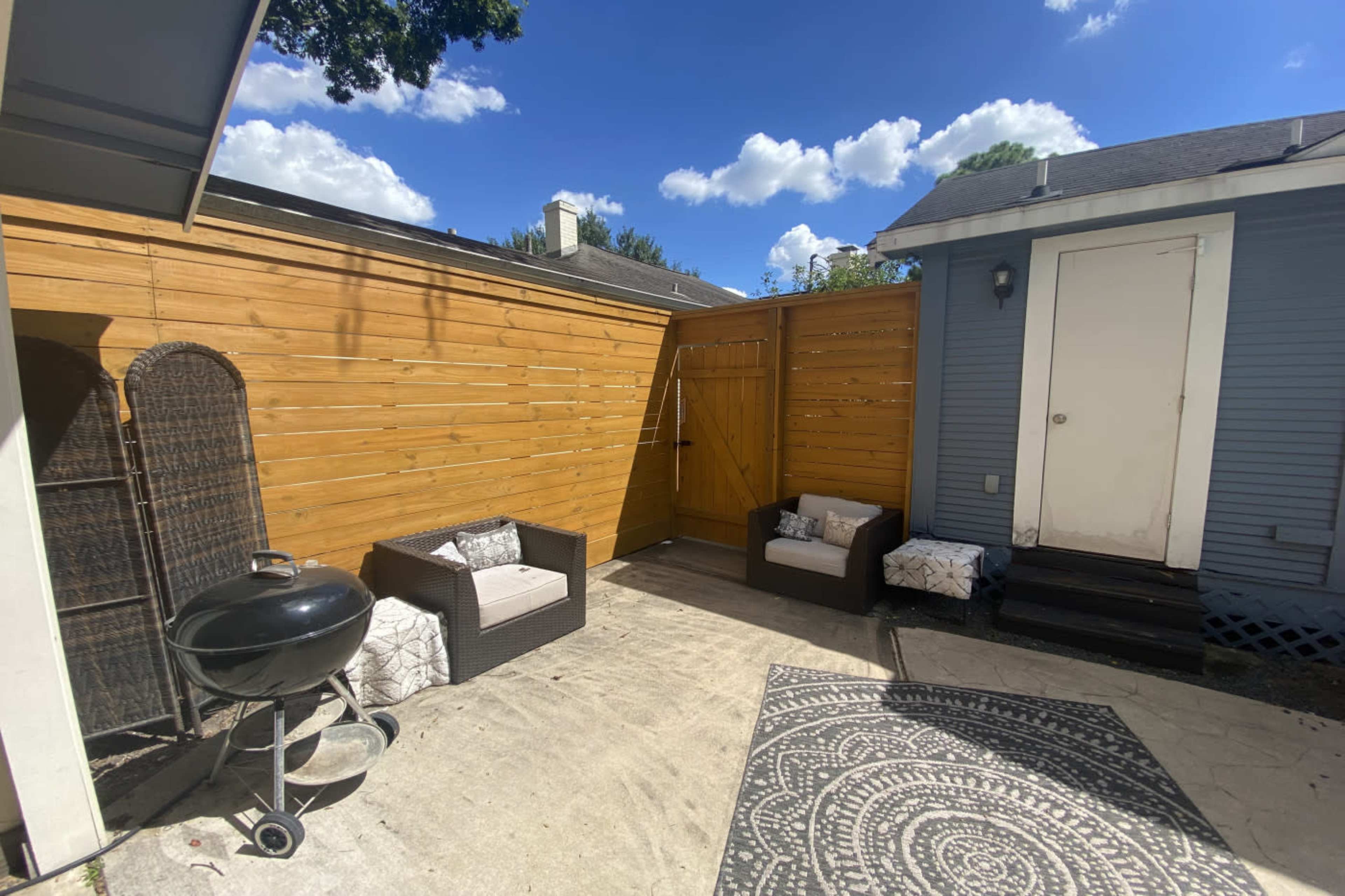 A small outdoor patio area featuring a wooden fence, two seating arrangements, a charcoal grill, and a decorative rug.